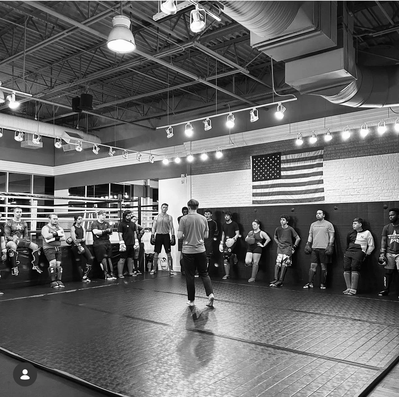 A group of people inside a boxing gym listening to a coach. The gym has an American flag on the wall, boxing gloves, and punching bags visible, with overhead lighting and exposed ceiling ductwork.