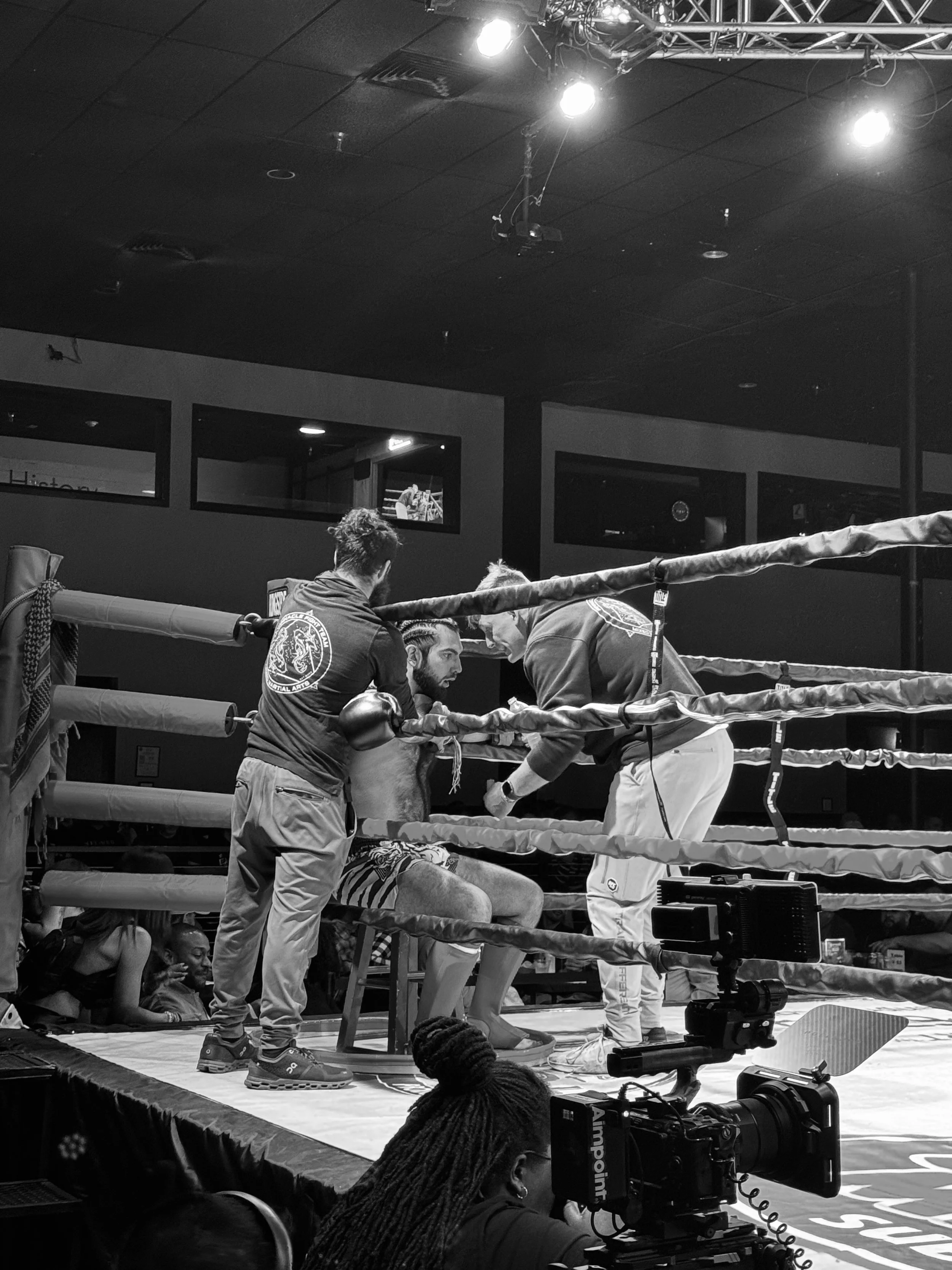 Boxers in a ring during a match, medics attending to an injured boxer on the ground, camera operators capturing the scene, and spectators in the background.