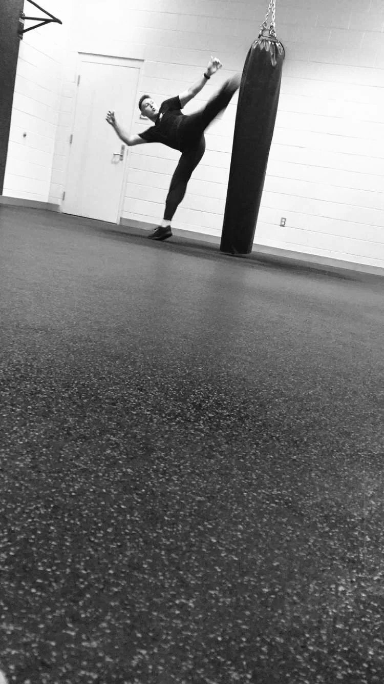A person practicing martial arts kicks a punching bag in a gym with a plain wall and closed door in the background.