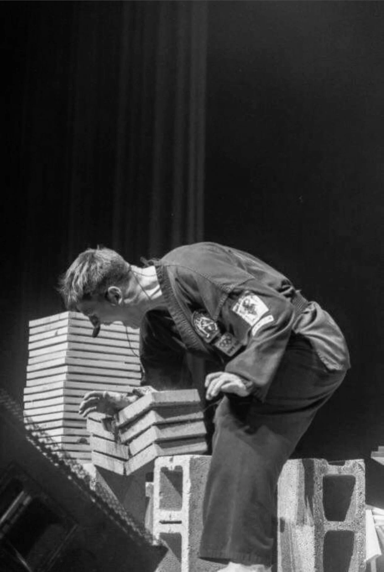 A person with short hair and glasses, dressed in a martial arts gi with patches, is kneeling and arranging or stacking bricks on a small table.