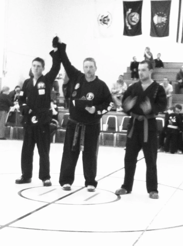 Black and white photo of a martial arts competition, with a referee holding a winner's hand up on a martial arts mat. Two martial artists stand nearby, one clapping. Spectators are seated on bleachers in the background.