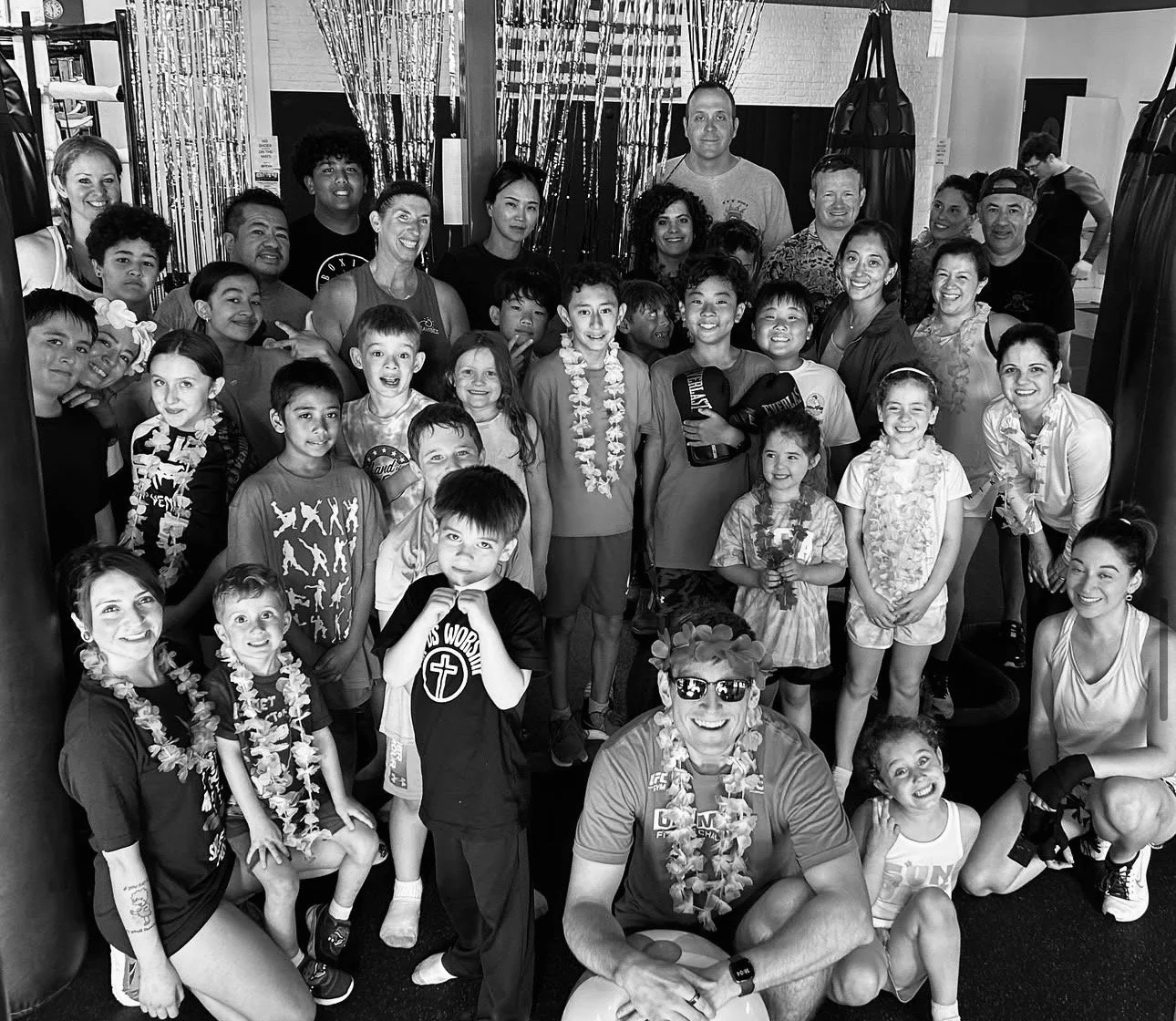 A large group of children and adults gathered together, smiling for a group photo in a gym or fitness center, with some wearing floral leis and casual athletic clothing.