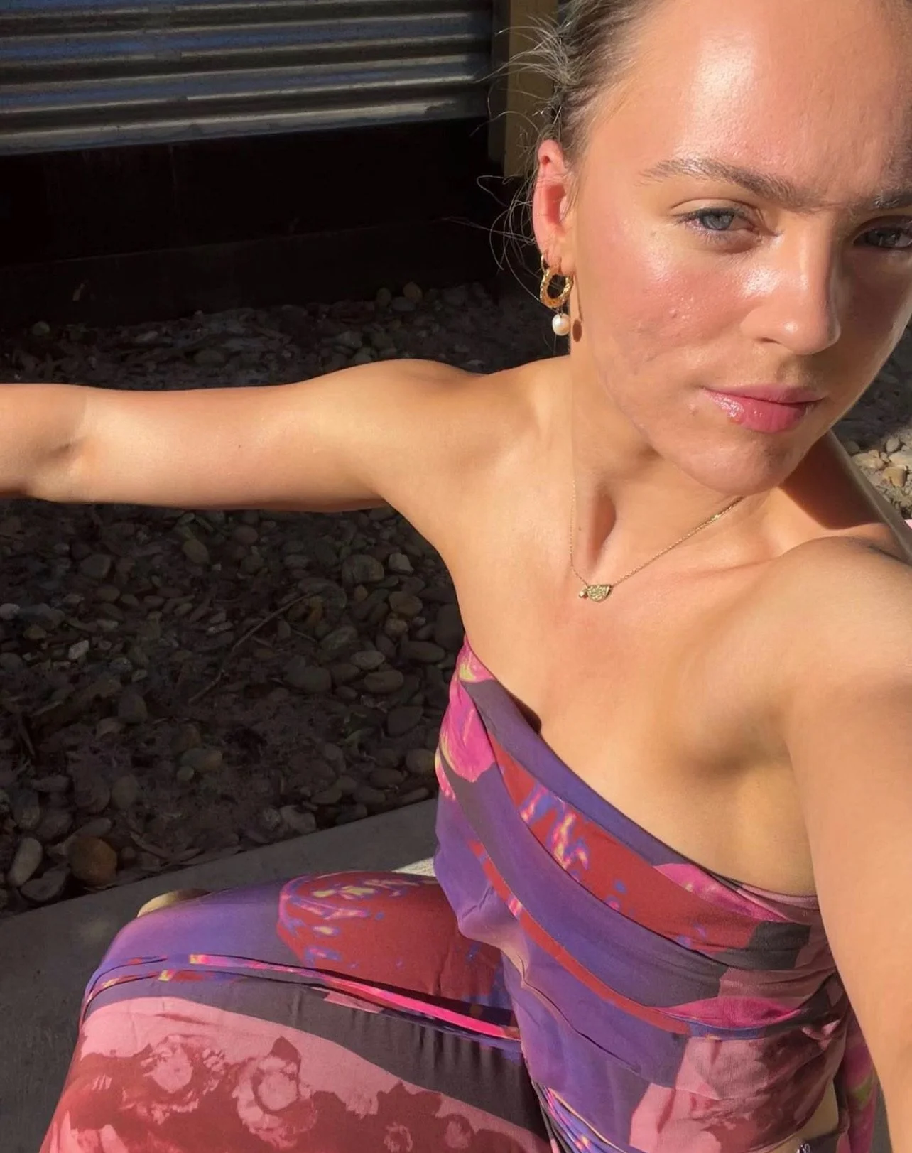A woman taking a selfie outdoors, wearing colorful clothing, earrings, a necklace, and has makeup on with sunlight on her face.