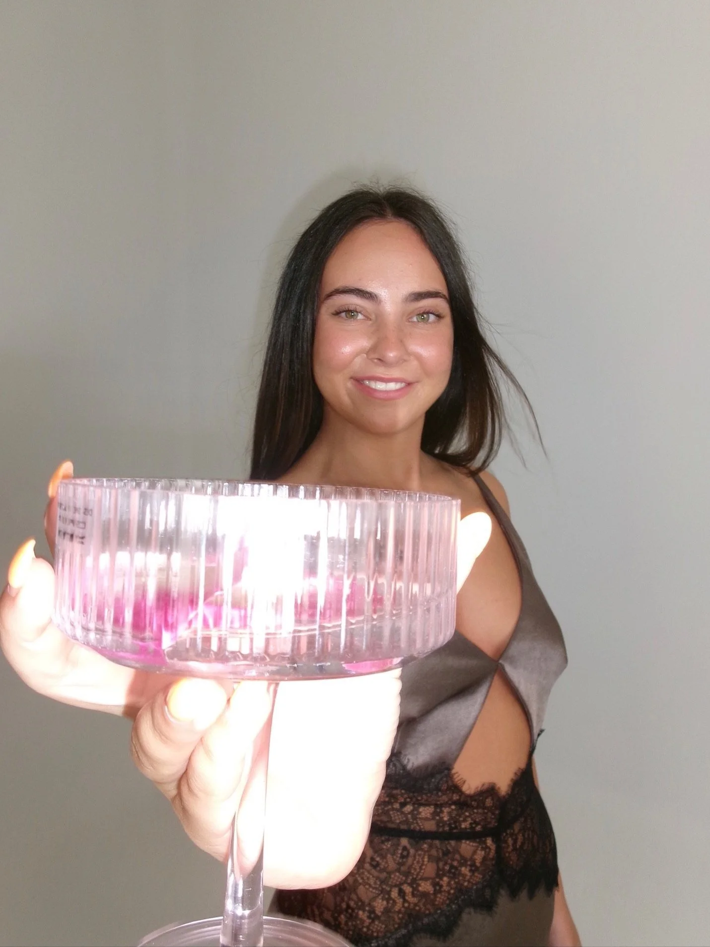 A woman with long dark hair smiling, wearing a sheer black lace and satin dress, holds a pink glass bowl towards the camera, with a plain, light-colored background.