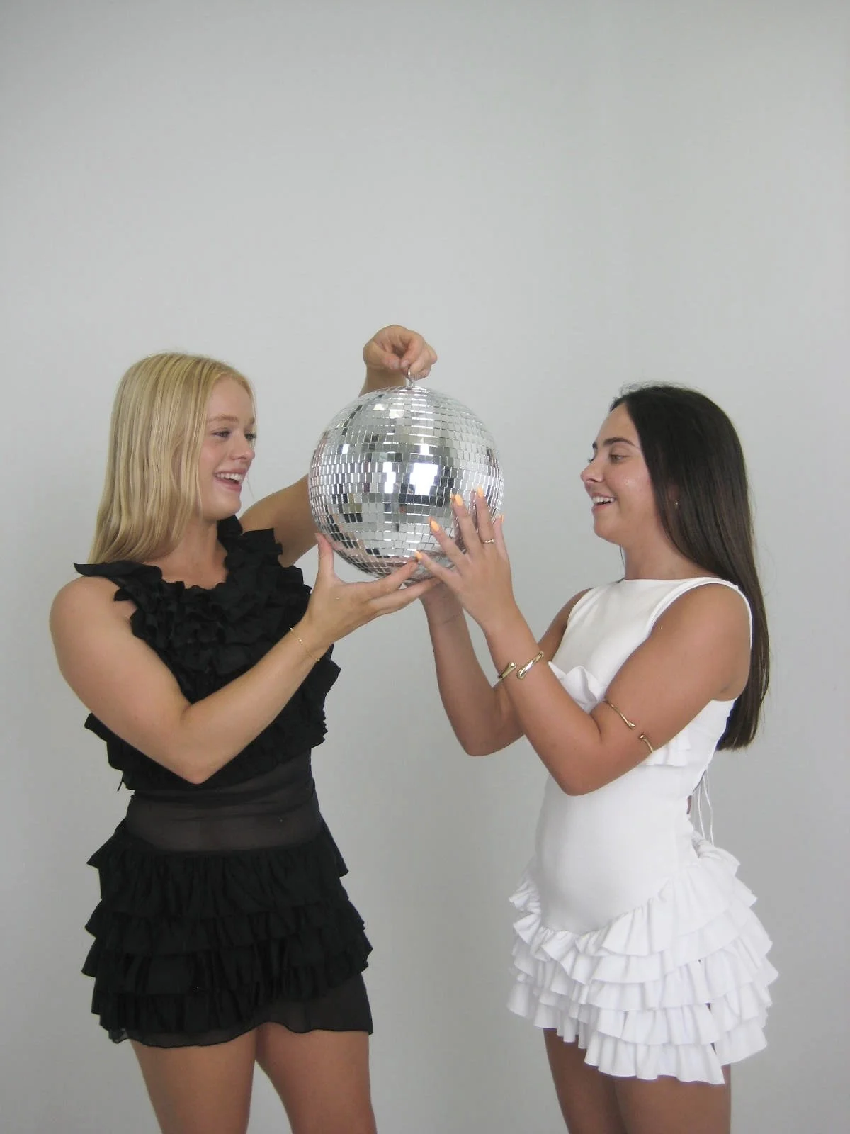 Two young women smiling and holding a shiny disco ball together against a plain white background.