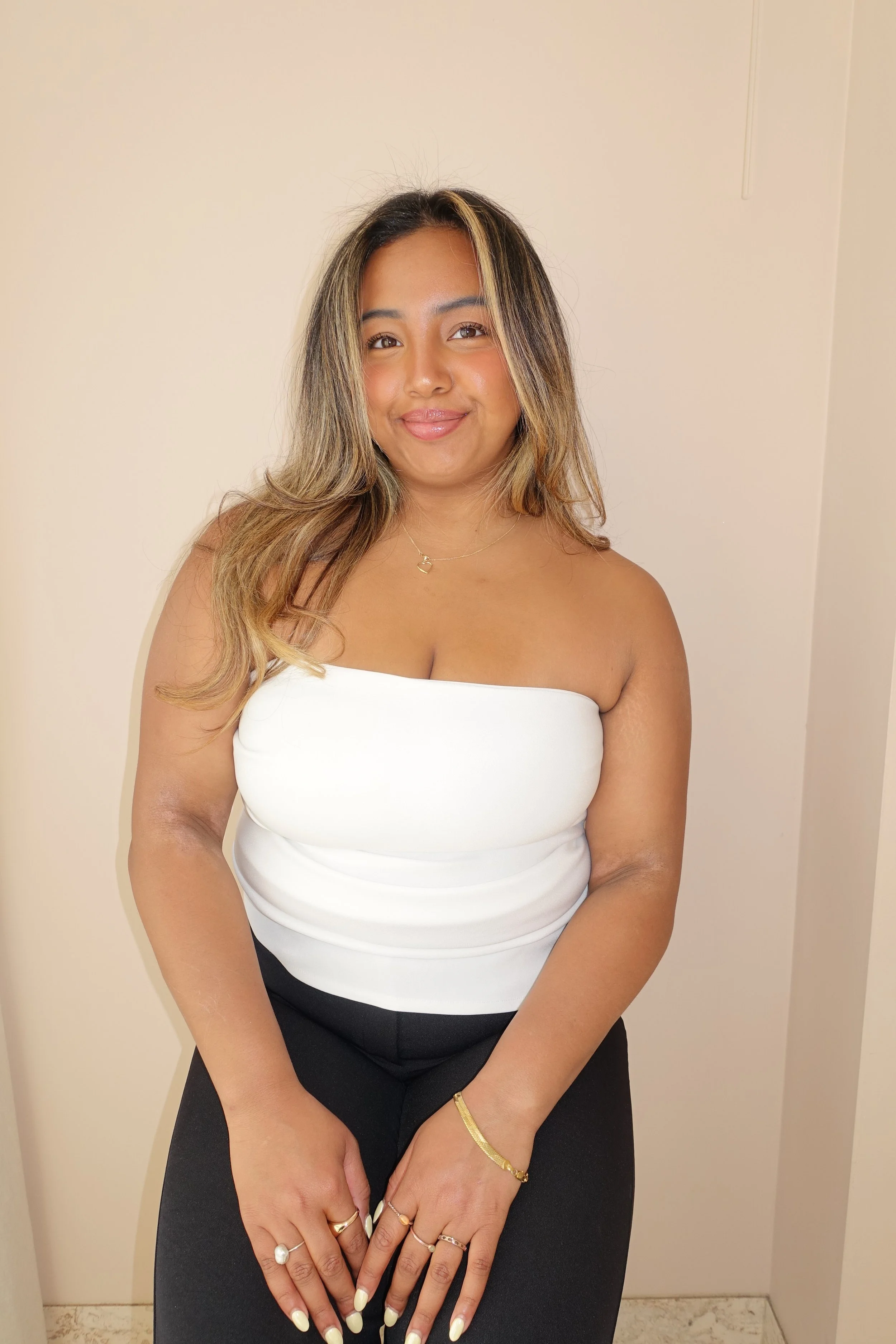 A woman with shoulder-length hair, wearing a white strapless top and black pants, smiling with hands clasped in front, decorated with rings and bracelets, standing against a plain wall in an indoor setting.
