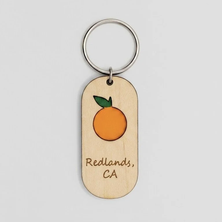 Wooden keychain with a painted orange fruit and green leaf, engraved with the text 'Redlands, CA'.