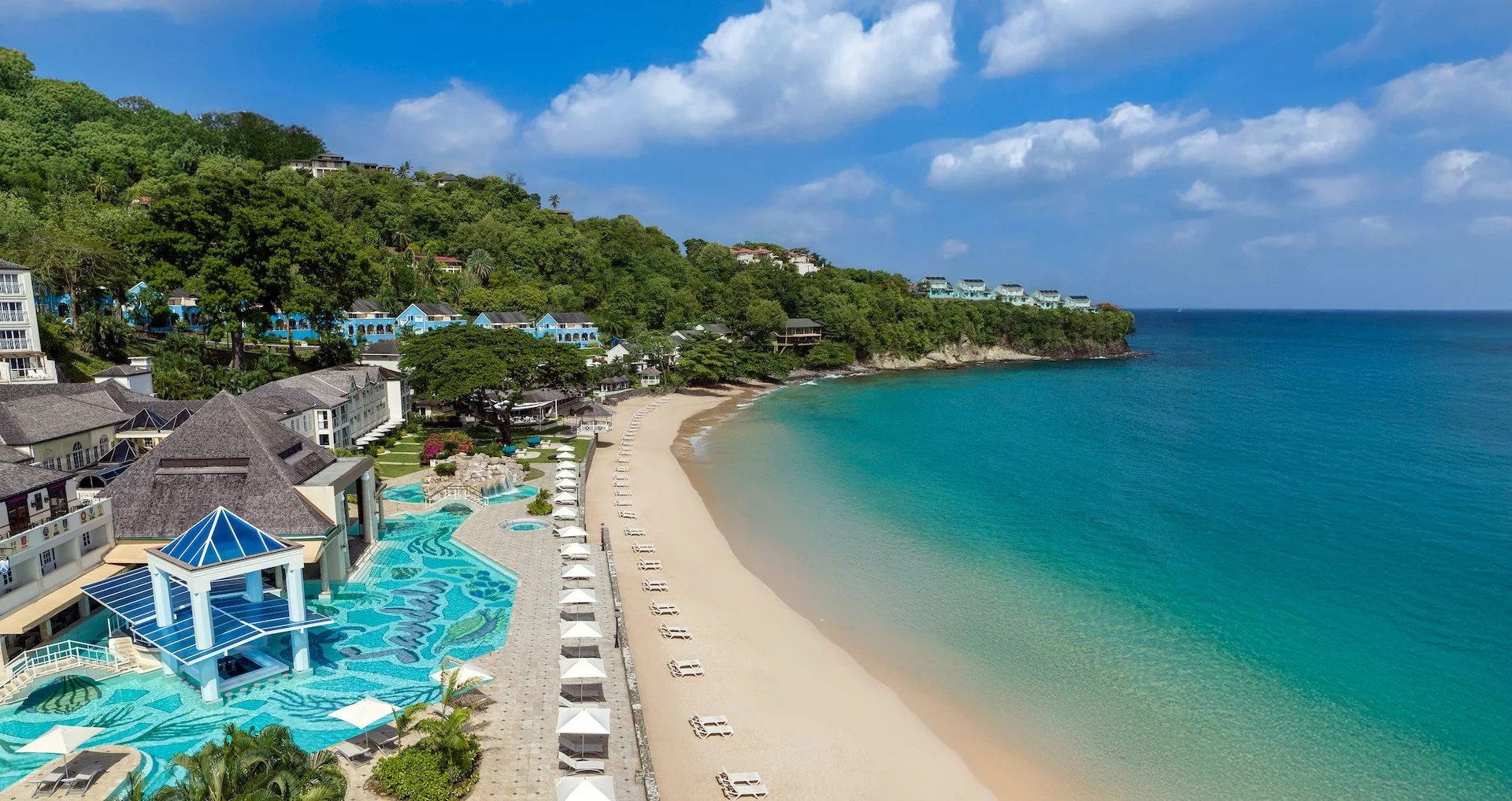 A beach with turquoise water, white sand, and lounge chairs with umbrellas. There are lush green hills with houses and resorts overlooking the beach. A resort pool area with a blue-roofed pavilion is visible in the foreground.