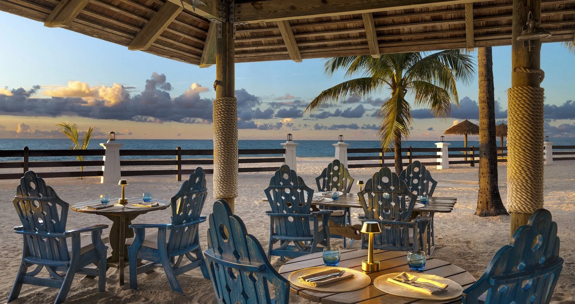 Outdoor beachfront restaurant with wooden tables and blue chairs under a thatched roof, overlooking the ocean at sunset with palm trees and clouds in the sky.