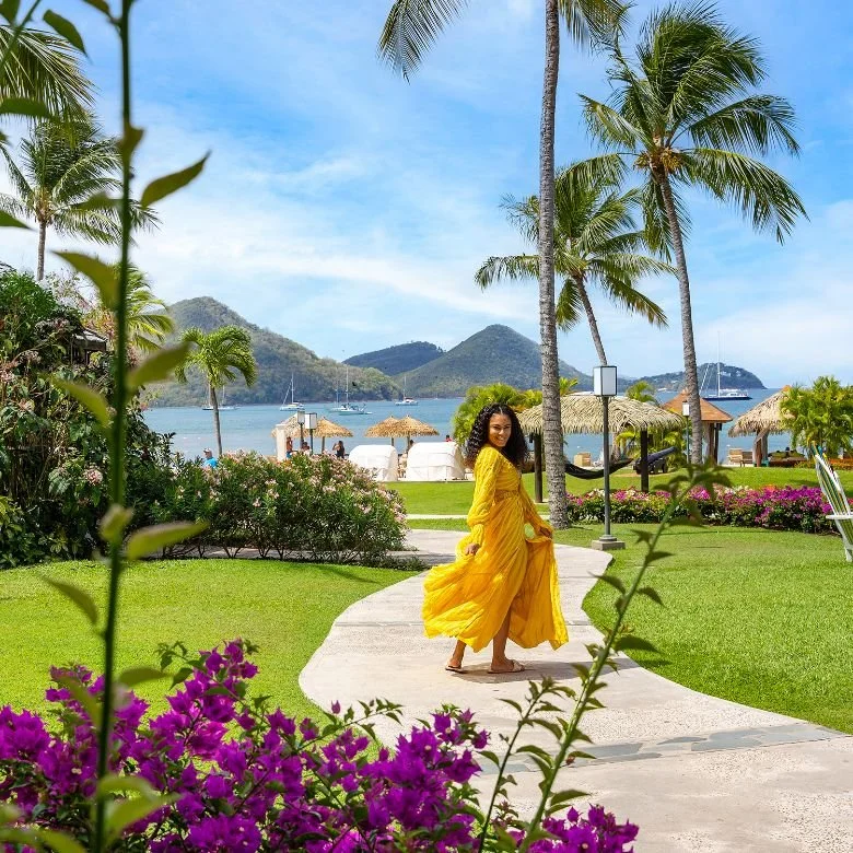 Woman in a yellow dress walking on a curved pathway in a lush tropical garden with palm trees, flowering bushes, and a view of the ocean and boats in the background, under a partly cloudy sky.