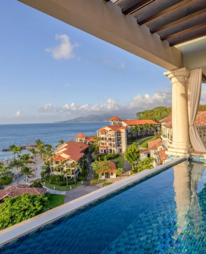 View of a resort with outdoor pool, palm trees, and multiple buildings with red roofs overlooking the ocean and mountains in the distance.