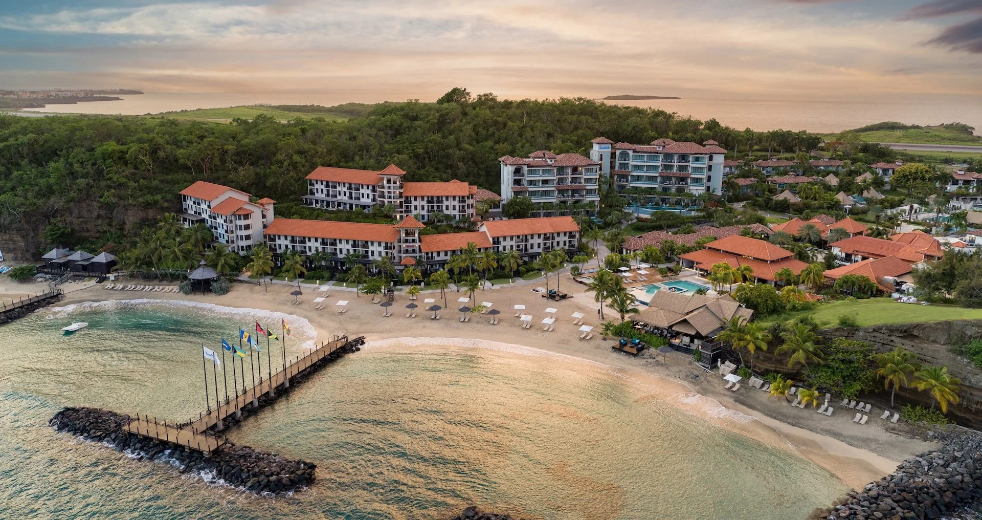Aerial view of a beachfront resort with multiple buildings, a sandy beach with lounge chairs and umbrellas, a pier with flags, and lush greenery near the ocean under a cloudy sky.