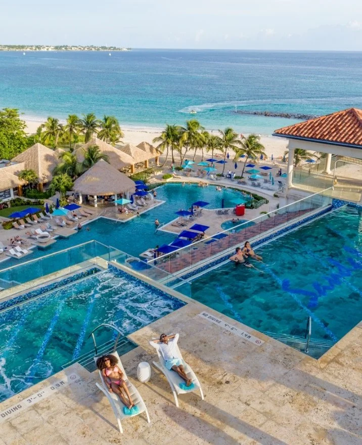 A view of a luxurious beachside resort with multiple pools, including a hot tub and lazy river, surrounded by lounge chairs, umbrellas, and palm trees, overlooking a sandy beach and ocean.
