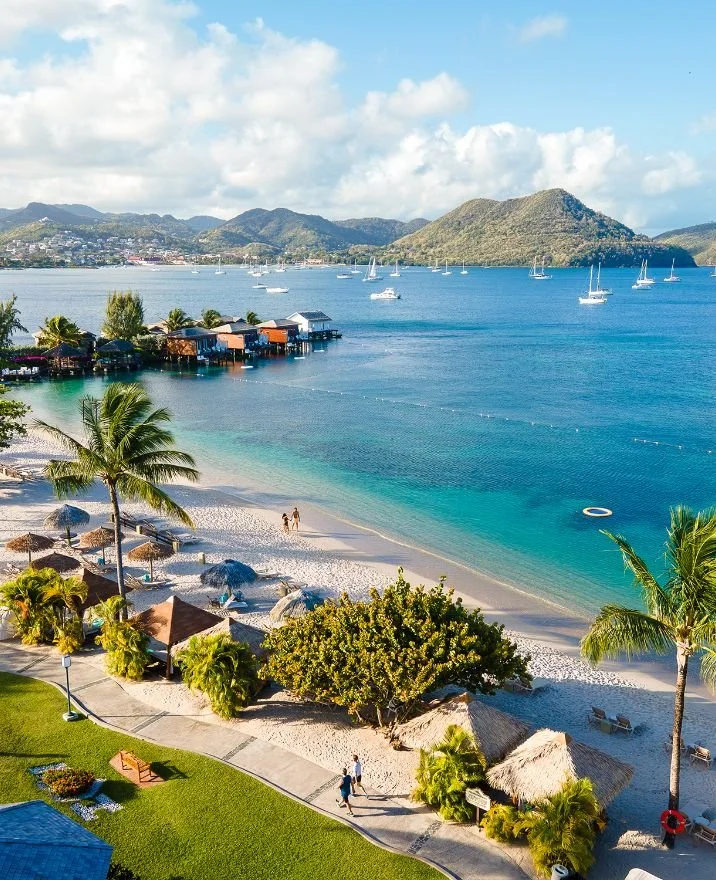 A tropical beach with white sand, palm trees, and umbrellas, overlooking blue ocean waters with sailboats and distant mountains.