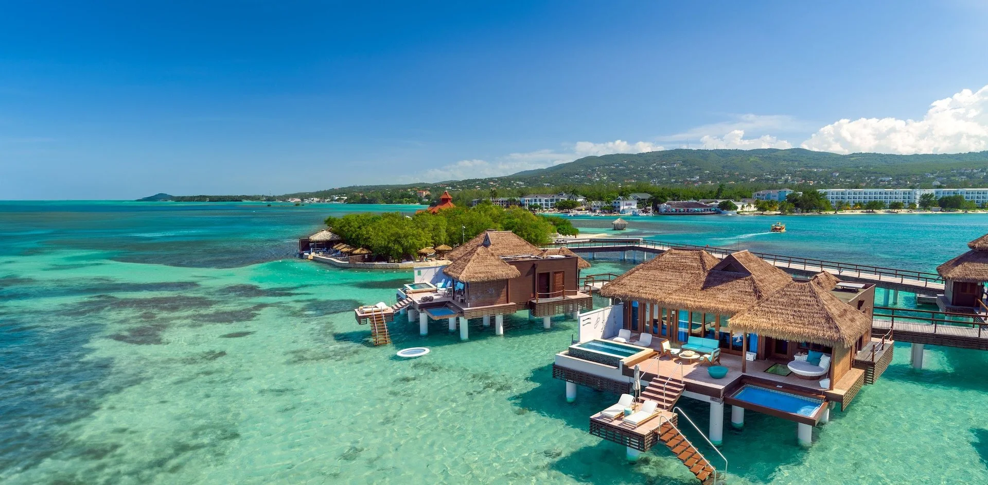 Overwater bungalows on clear turquoise sea with greenery and hills in the background.