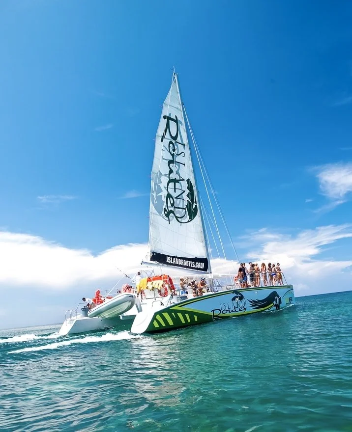 A large catamaran sailboat with a blue and green design named Island Routes, carrying multiple people on the deck, sailing on clear turquoise water under a bright blue sky.
