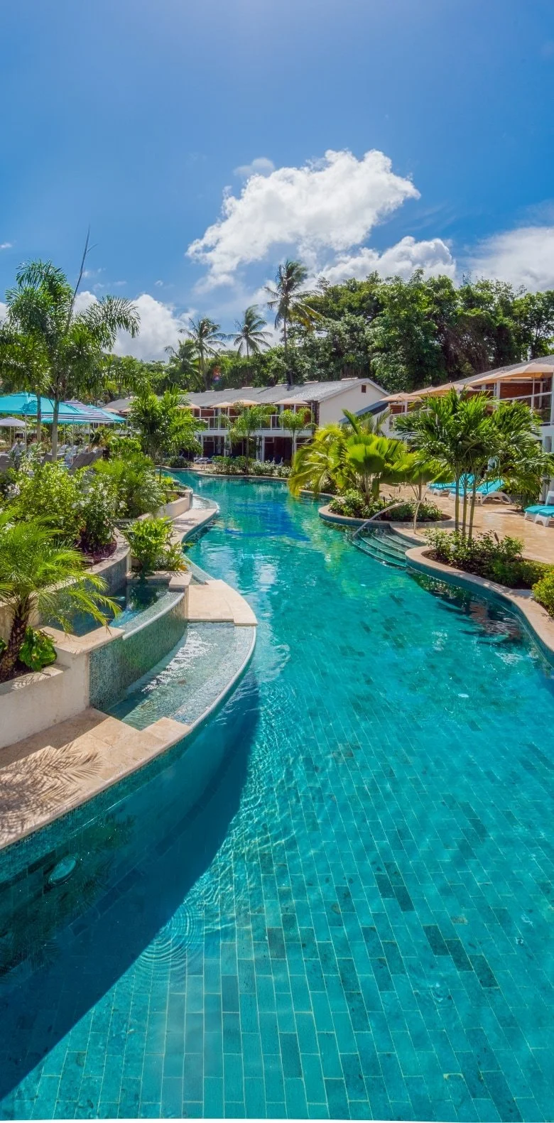 Resort swimming pool surrounded by tropical plants, lounge chairs, and umbrellas, with a blue sky and white clouds overhead.