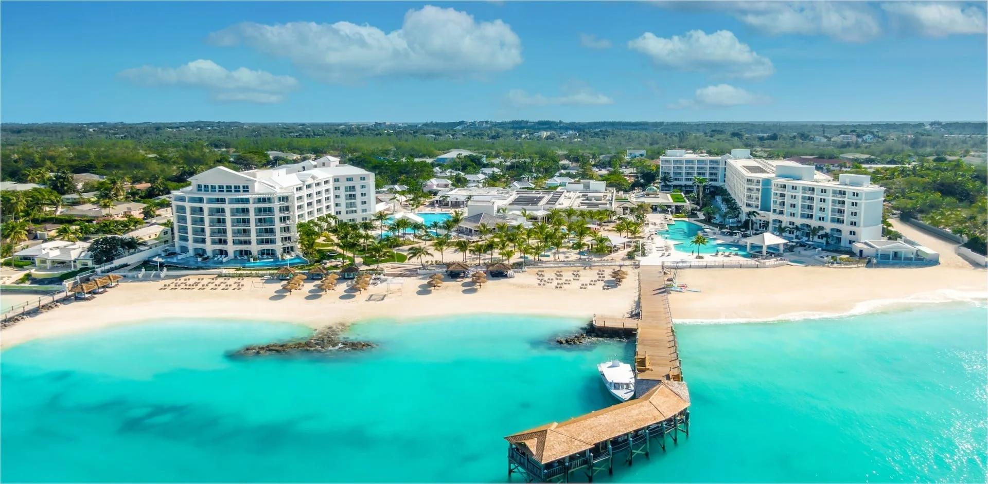 Aerial view of a beachfront resort with white buildings, a sandy beach with umbrellas, turquoise water, and a pier with a boat at the end.