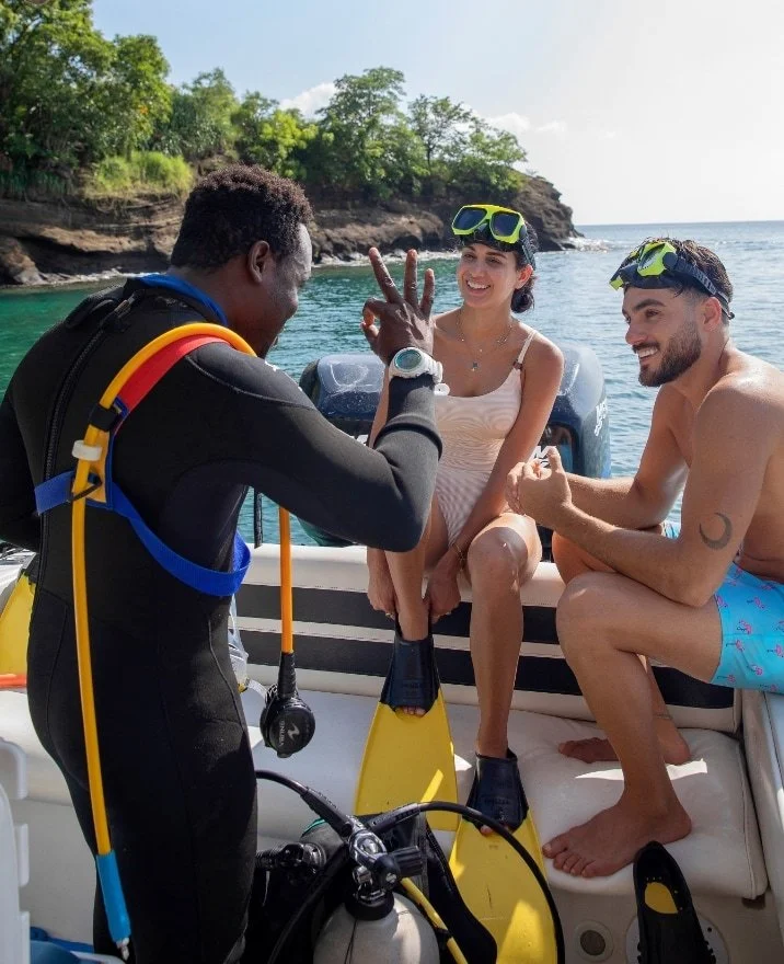 A scuba instructor in a black wetsuit shows two happy tourists, a woman in a beige dress and a man in blue swim trunks, on a boat near the coast with rocky cliffs and sea in the background.
