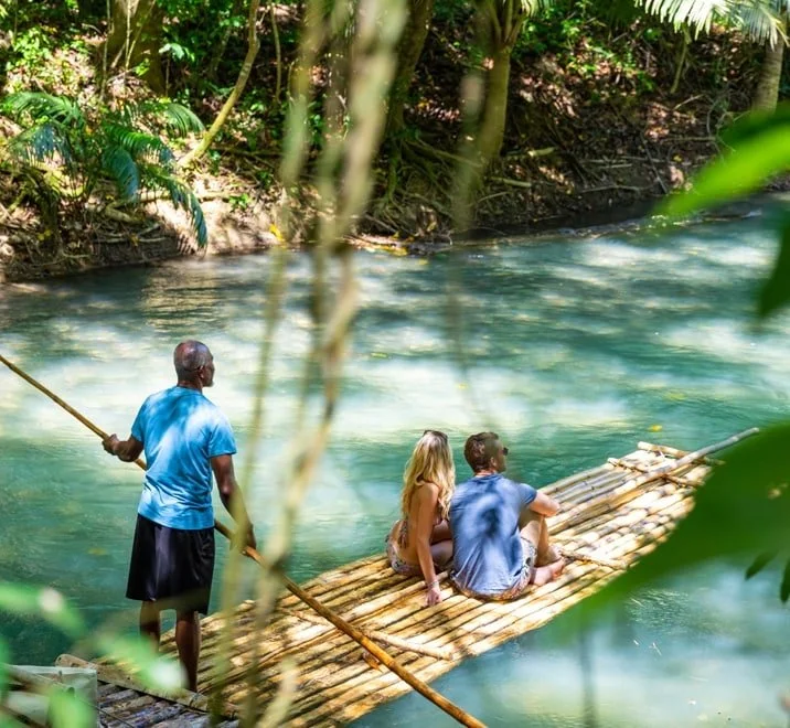 Two tourists sitting on a bamboo raft on a river in a lush tropical jungle, with a local guide standing nearby holding a pole.