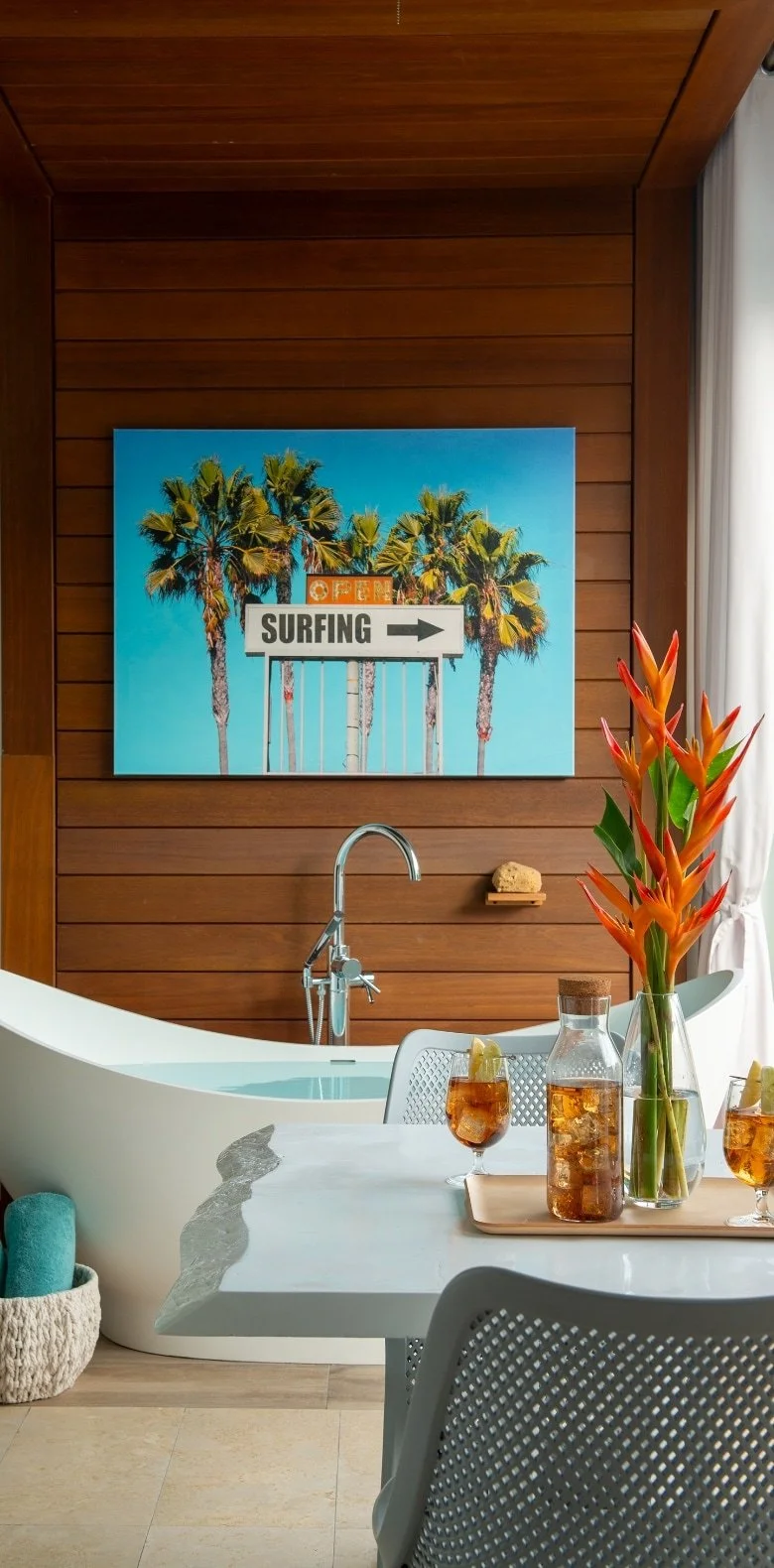 A bathroom with a wooden wall, a bathtub, a sink with a faucet, a basket with a towel, a table with drinks and a vase with tropical flowers, and a large wall art of palm trees and a surf sign.