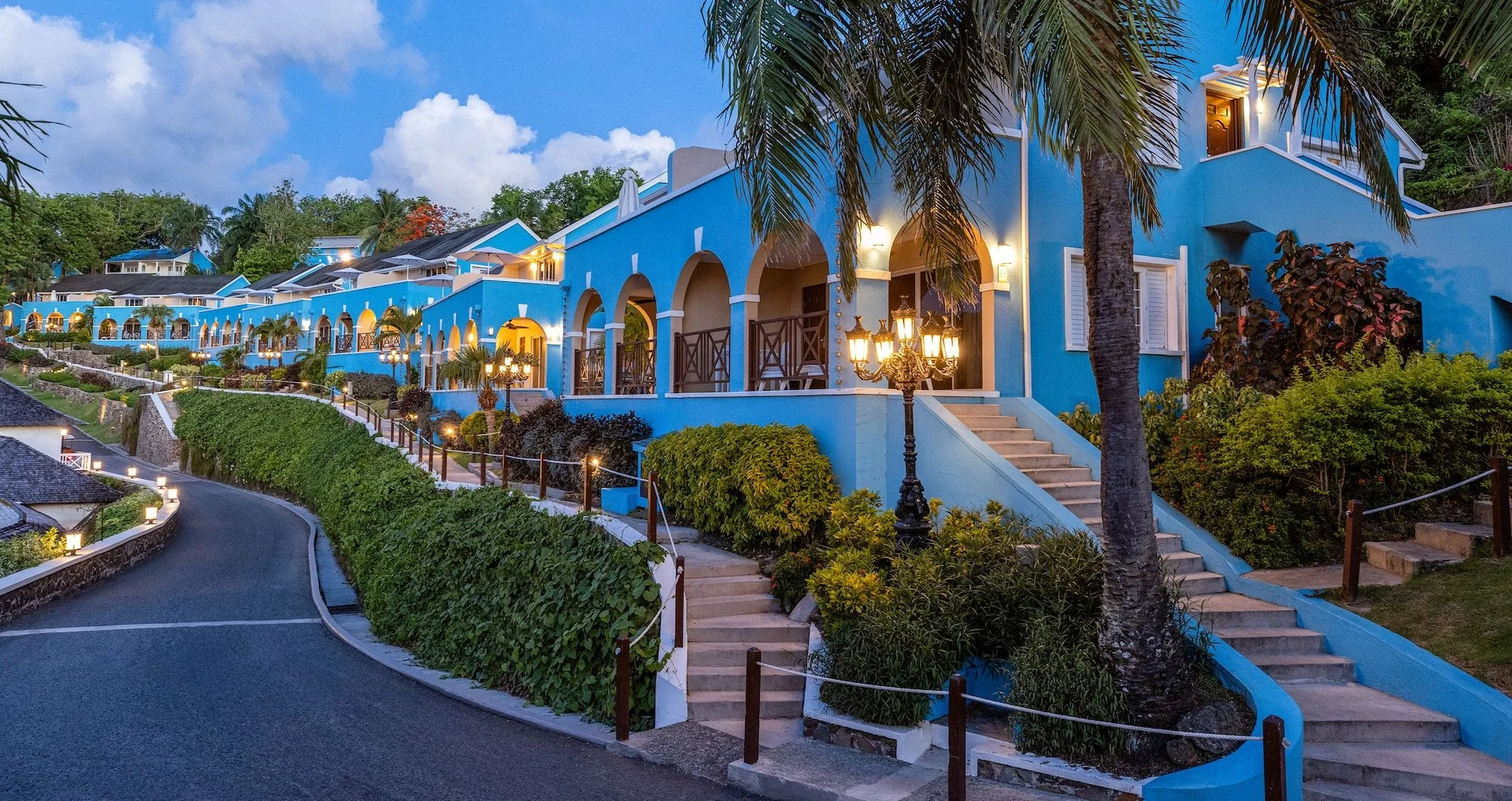 A series of brightly lit blue Mediterranean-style villas on a hillside with palm trees and lush greenery, evening setting with twilight sky.