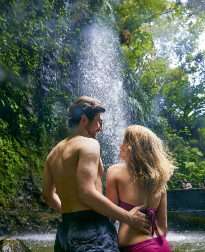 A couple with topless upper bodies in front of a waterfall surrounded by lush greenery.