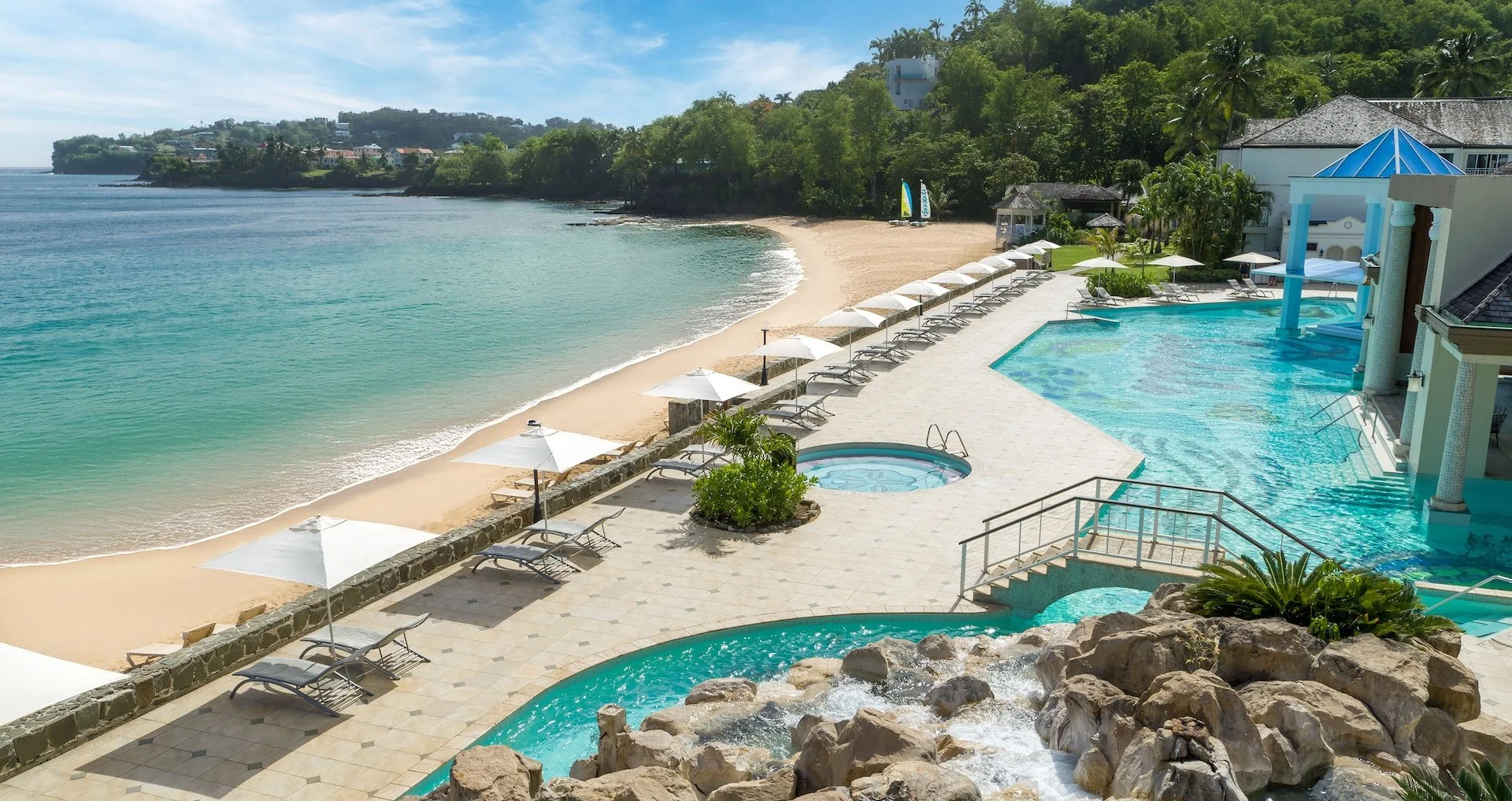 Resort pool area overlooking a sandy beach and ocean with greenery in the background. White lounge chairs and umbrellas are lined along the poolside.