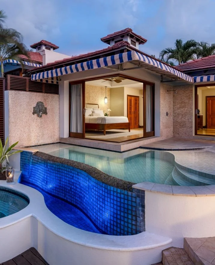 View of a luxurious backyard with a small pool, an open sliding door revealing a bedroom, and a covered outdoor area with striped blue and white awnings.