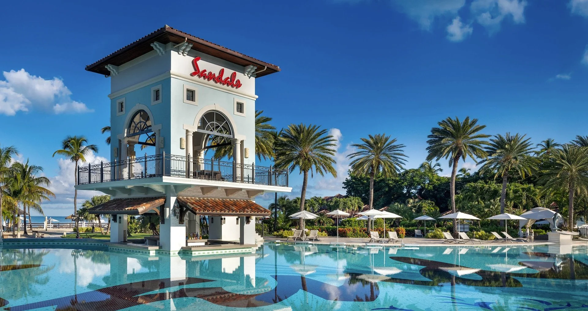 Luxury resort pool area with a white tower labeled "Sandals," surrounded by palm trees, lounge chairs, and umbrellas against a blue sky.