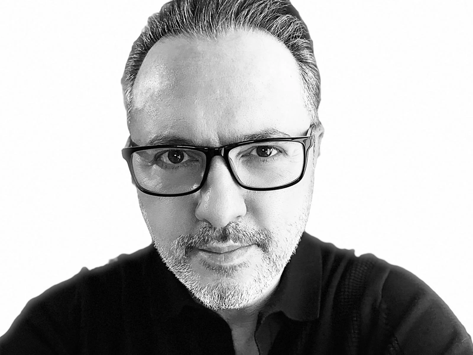 A black and white close-up selfie of a man with glasses and a beard, looking directly at the camera with a neutral expression.