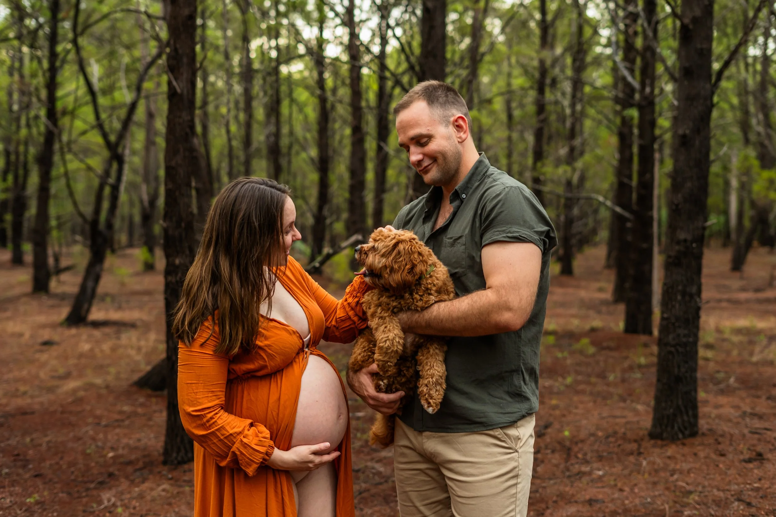 A pregnant woman with long brown hair wearing an orange dress and a man with short brown hair and facial hair, wearing a green shirt, are standing in a forest. The woman is touching her pregnant belly while the man is holding a small, curly-haired br