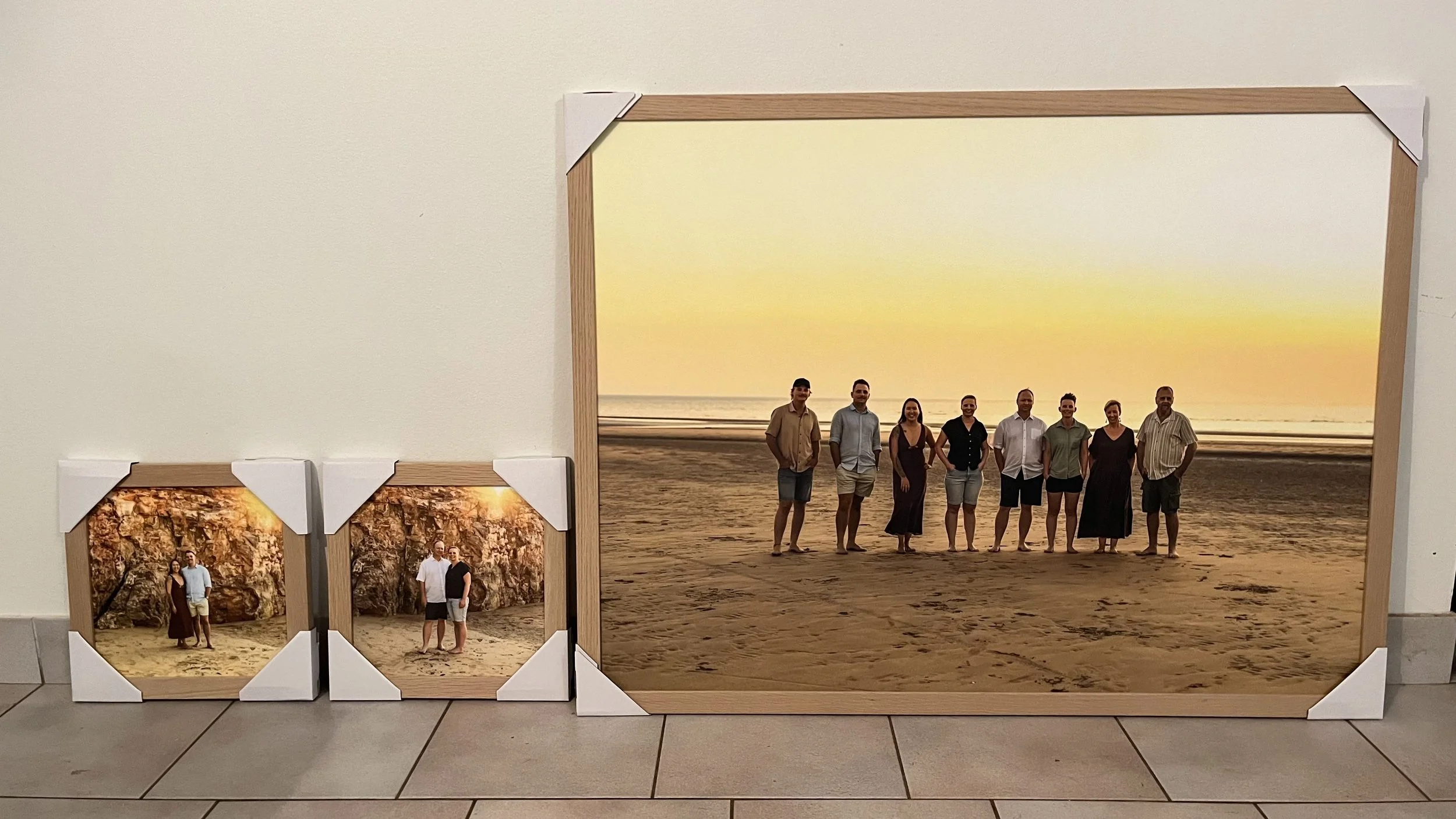 Three framed photographs of a group of people at the beach are leaning against a white wall on tiled flooring. The largest photo features nine people standing on the sand at sunset with the ocean in the background. The two smaller photos show the same group at a different location with rocks and trees.