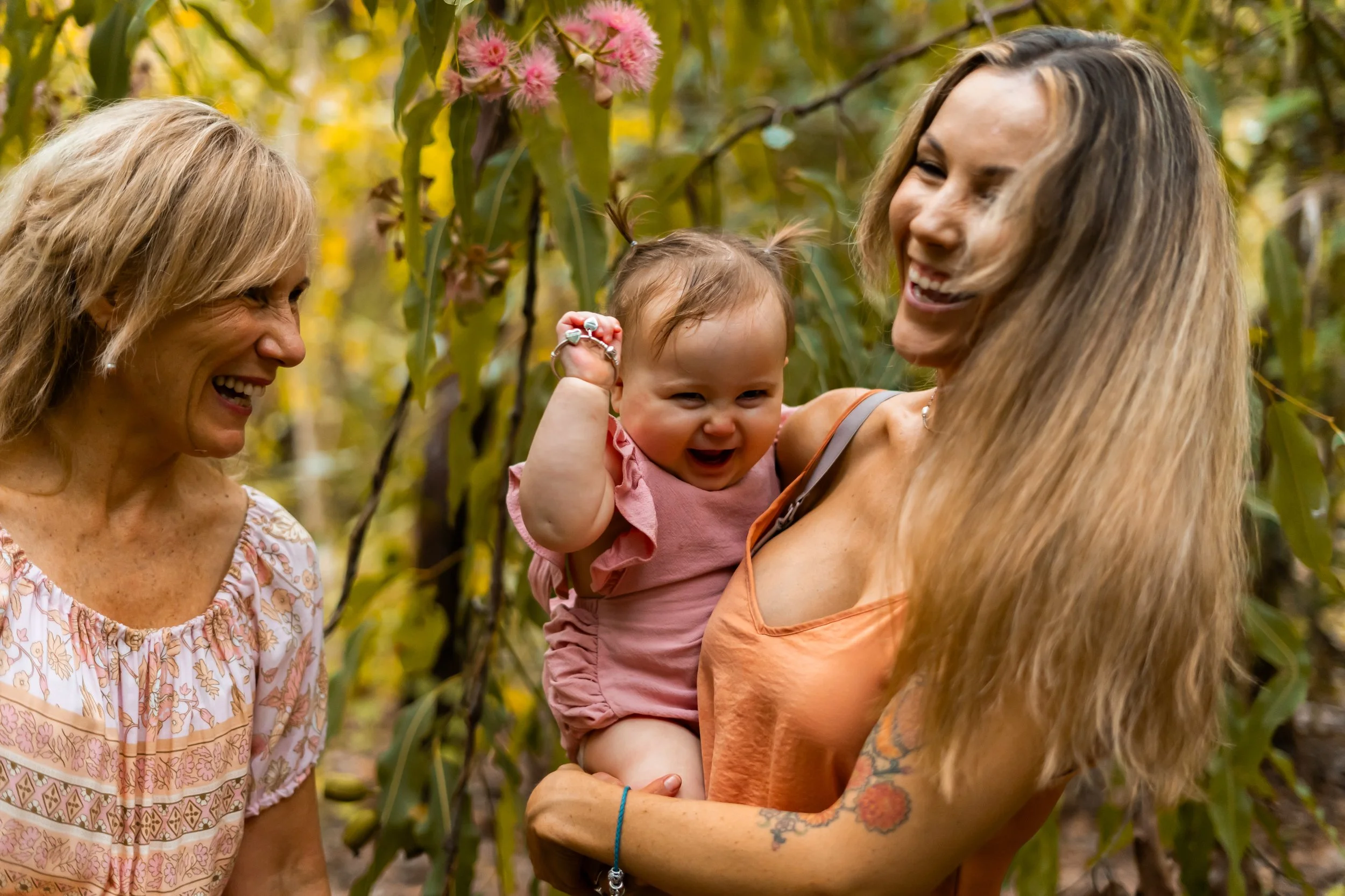 Two women and a baby girl outdoors surrounded by yellow-green foliage, smiling and laughing.