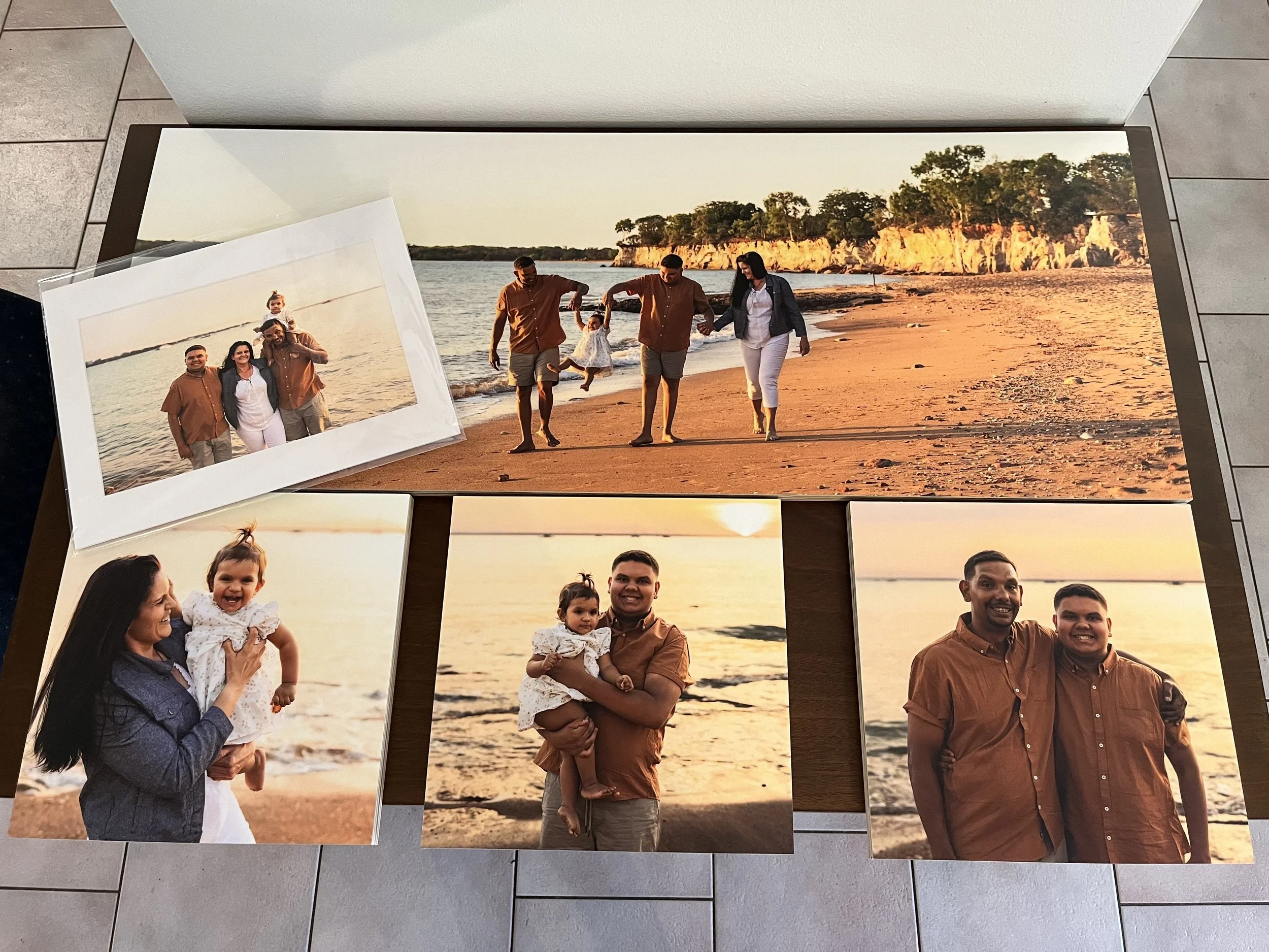 A collection of five printed photos on a wooden table, capturing a family at the beach during sunset. The photos include a group shot of four people, a family walking along the shoreline, a woman holding a smiling child, a man holding the same child, and two men standing together, all enjoying their time at the beach.
