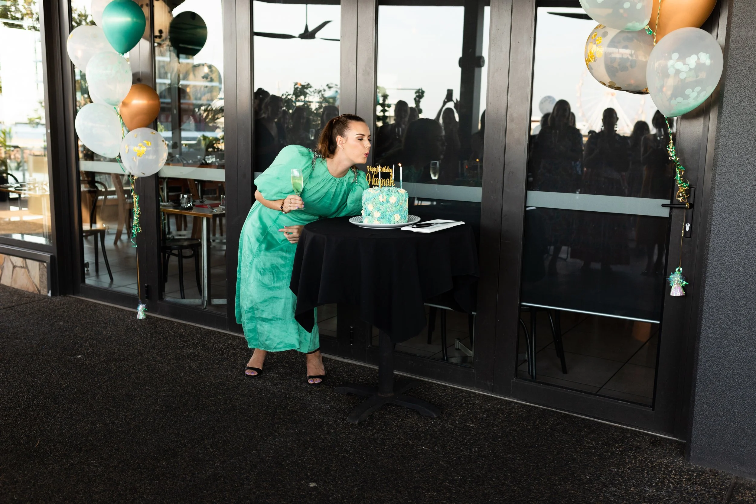 A young woman sits on the beach sand near the ocean at sunset, smiling and looking into the distance, wearing an off-shoulder green dress.