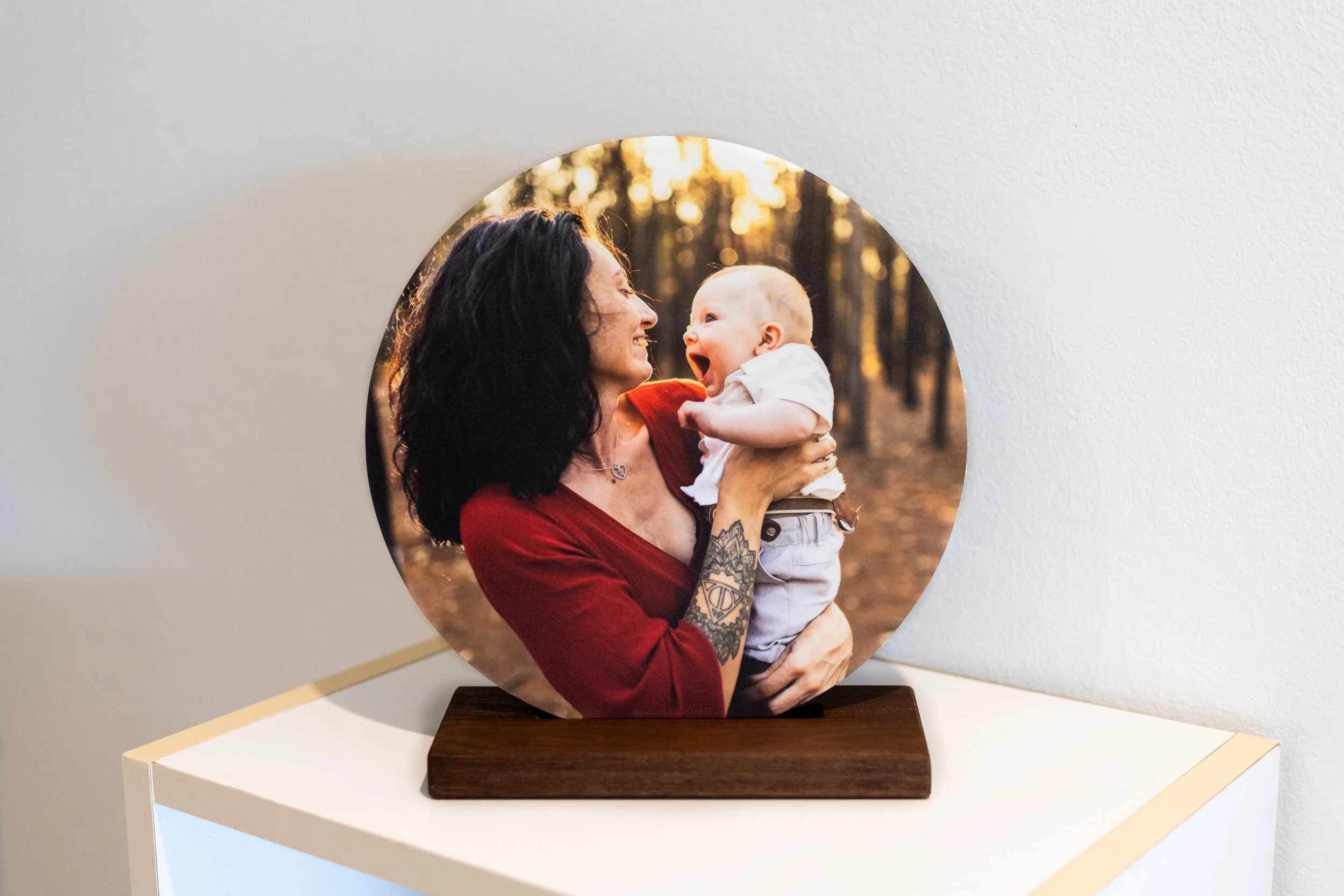 A circular photo of a woman with dark curly hair holding a laughing baby in a white shirt and beige shorts in a wooded park at sunset.