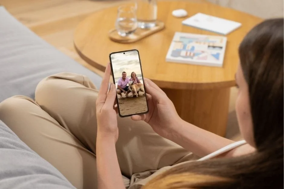 A woman sitting on a couch holds a smartphone, taking a photo of two people and a dog on the beach.