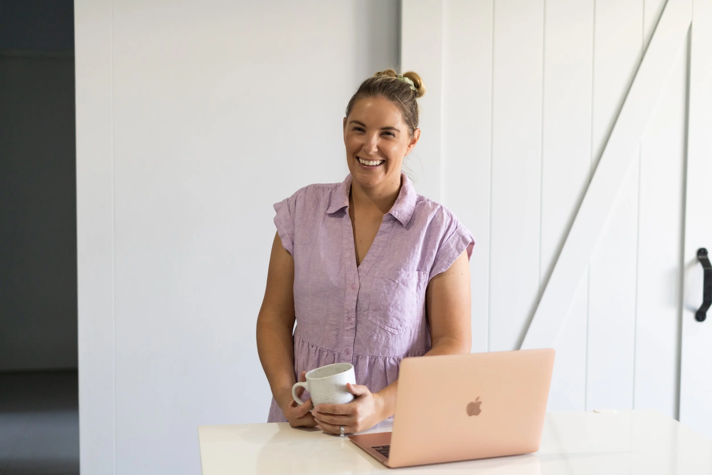 A smiling woman standing at a white table holding a mug, with a pink MacBook on the table in front of her, in a bright room with white walls.