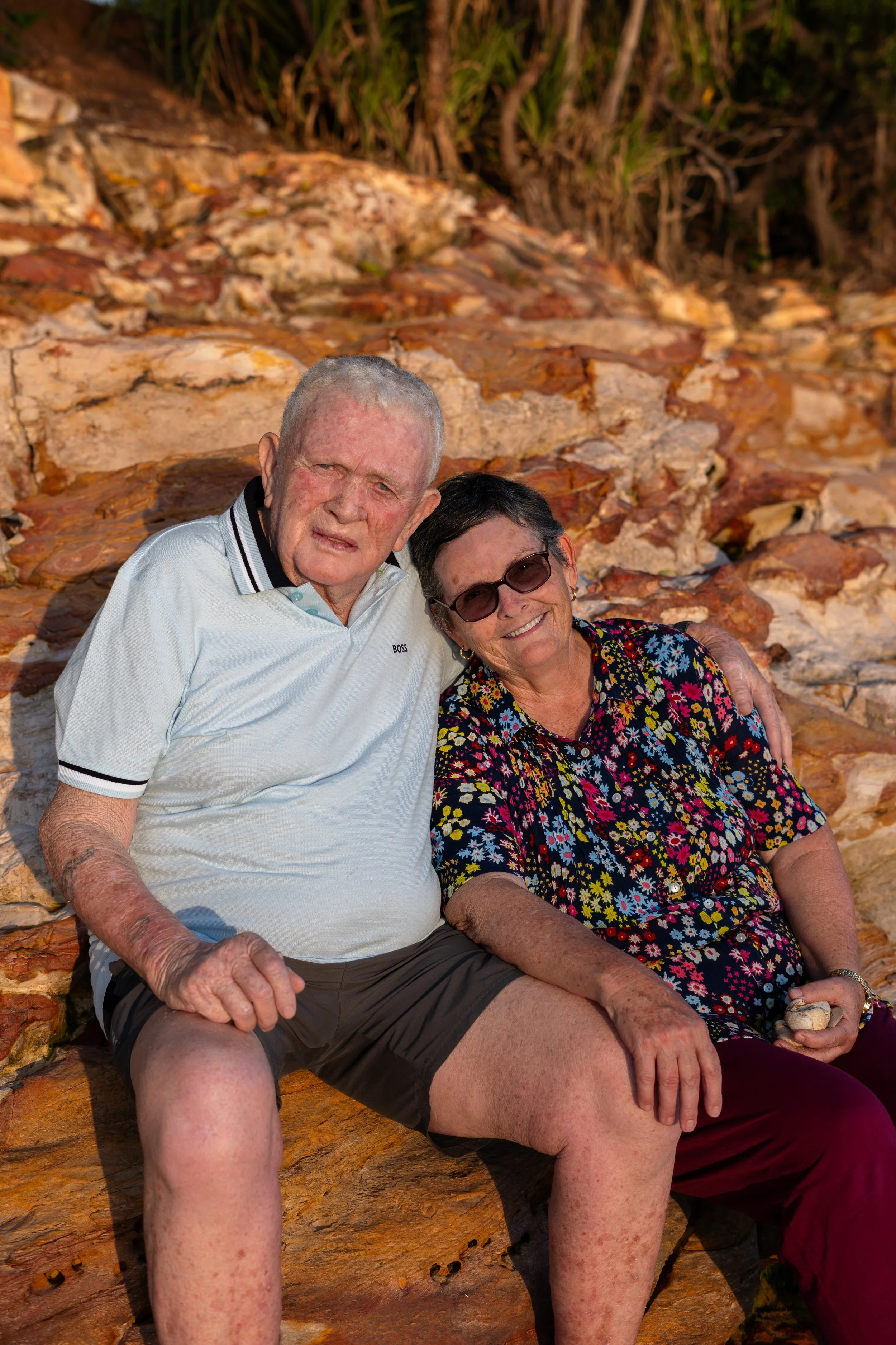An elderly man and woman sitting side by side on rocky terrain outdoors, smiling and enjoying each other's company.