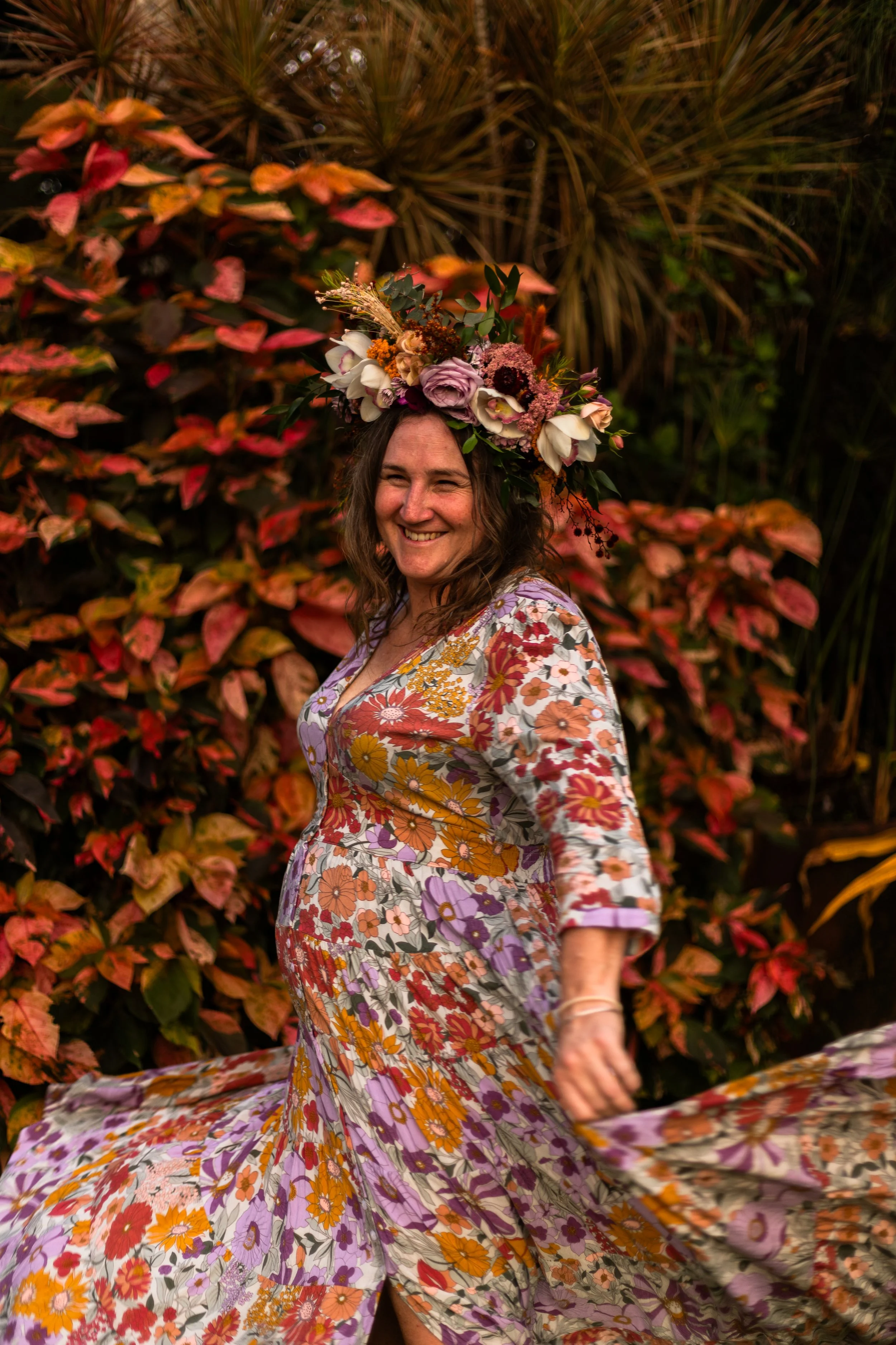 A woman in a colorful floral dress with a flower crown, smiling and twirling outdoors with red and green foliage in the background.
