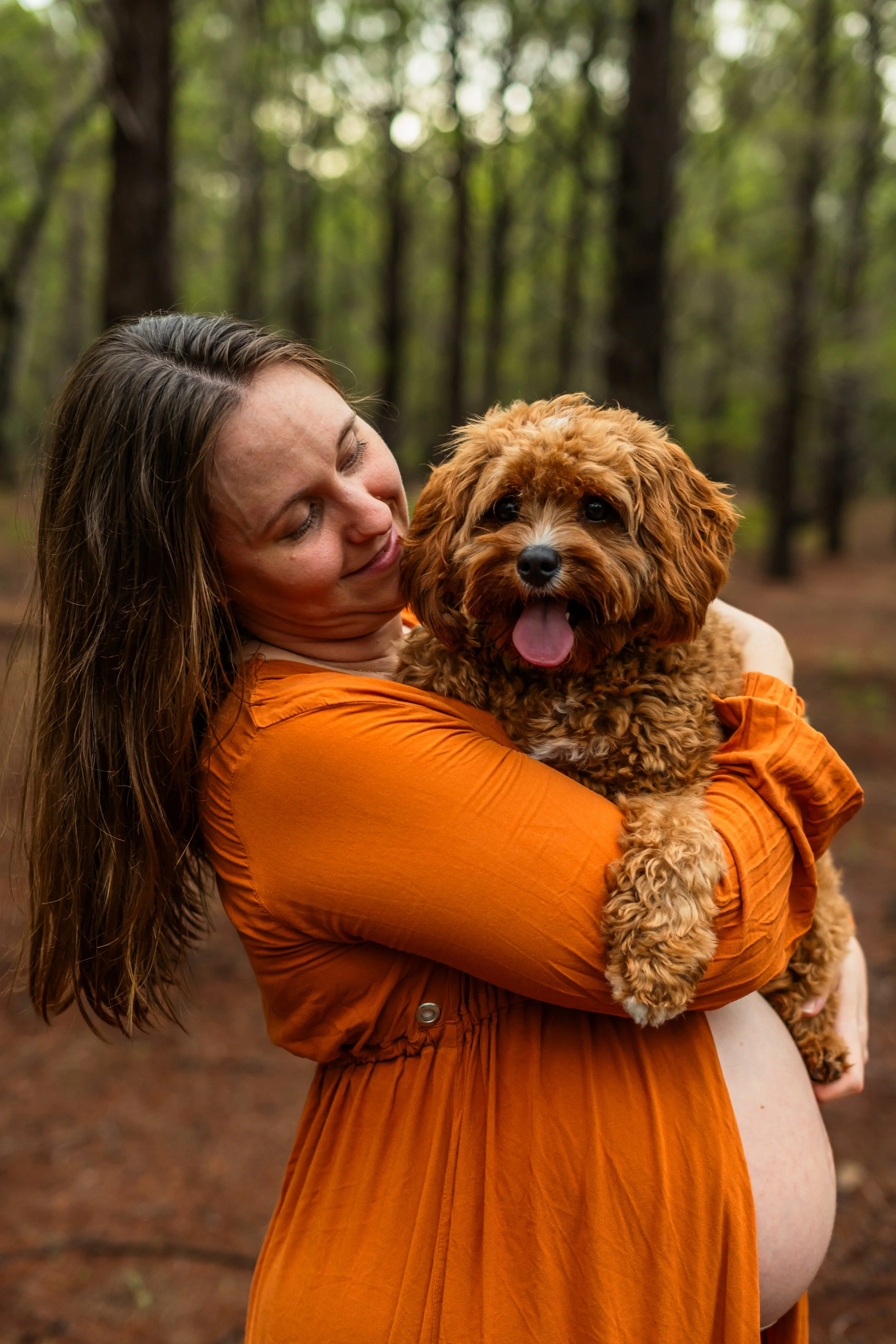 A woman with long hair and a bright orange dress is holding a curly-haired brown puppy with a white nose and paws, in a forested area.