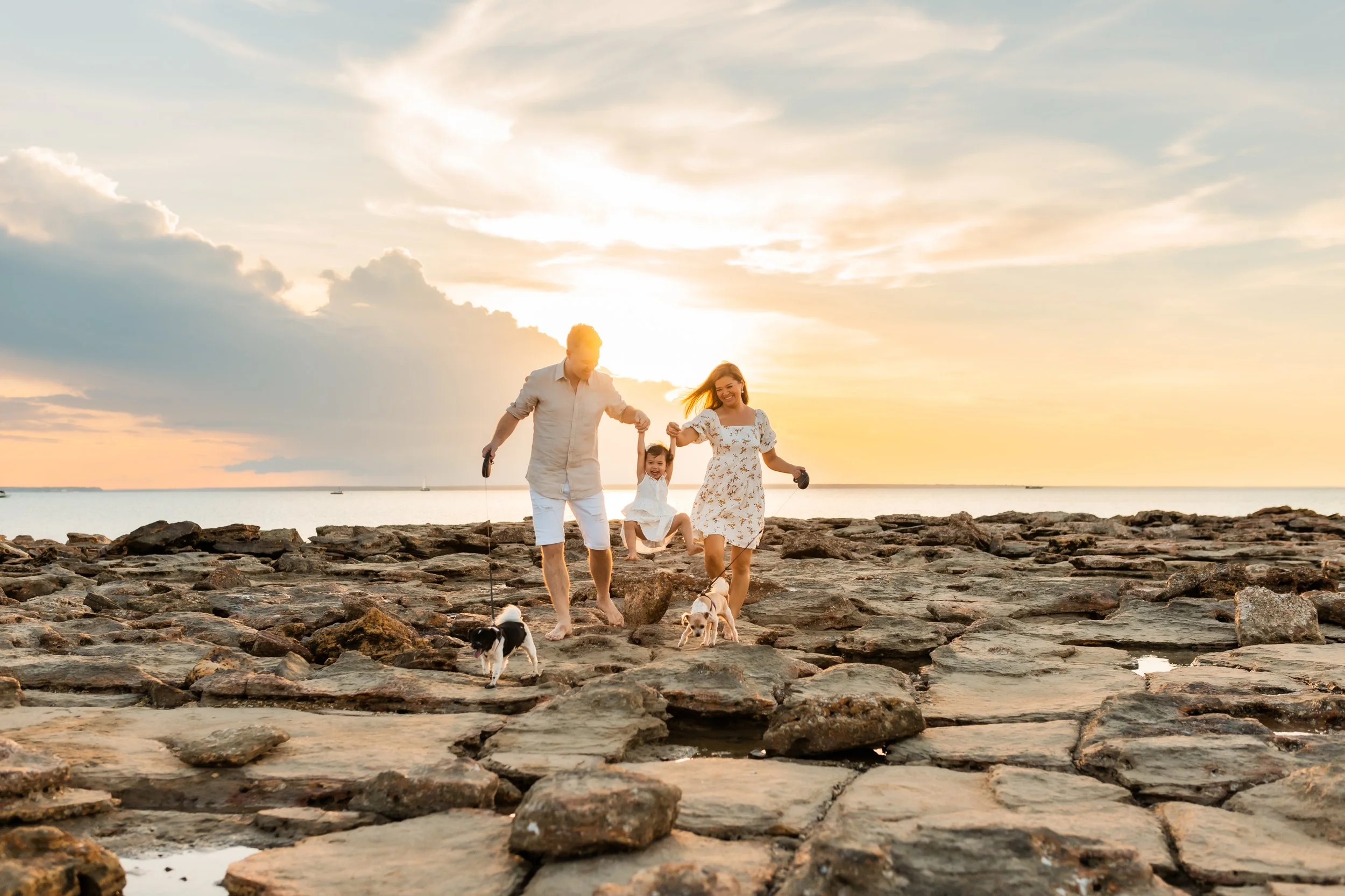 A family walking on a rocky beach at sunset, holding hands and playing with their dogs.
