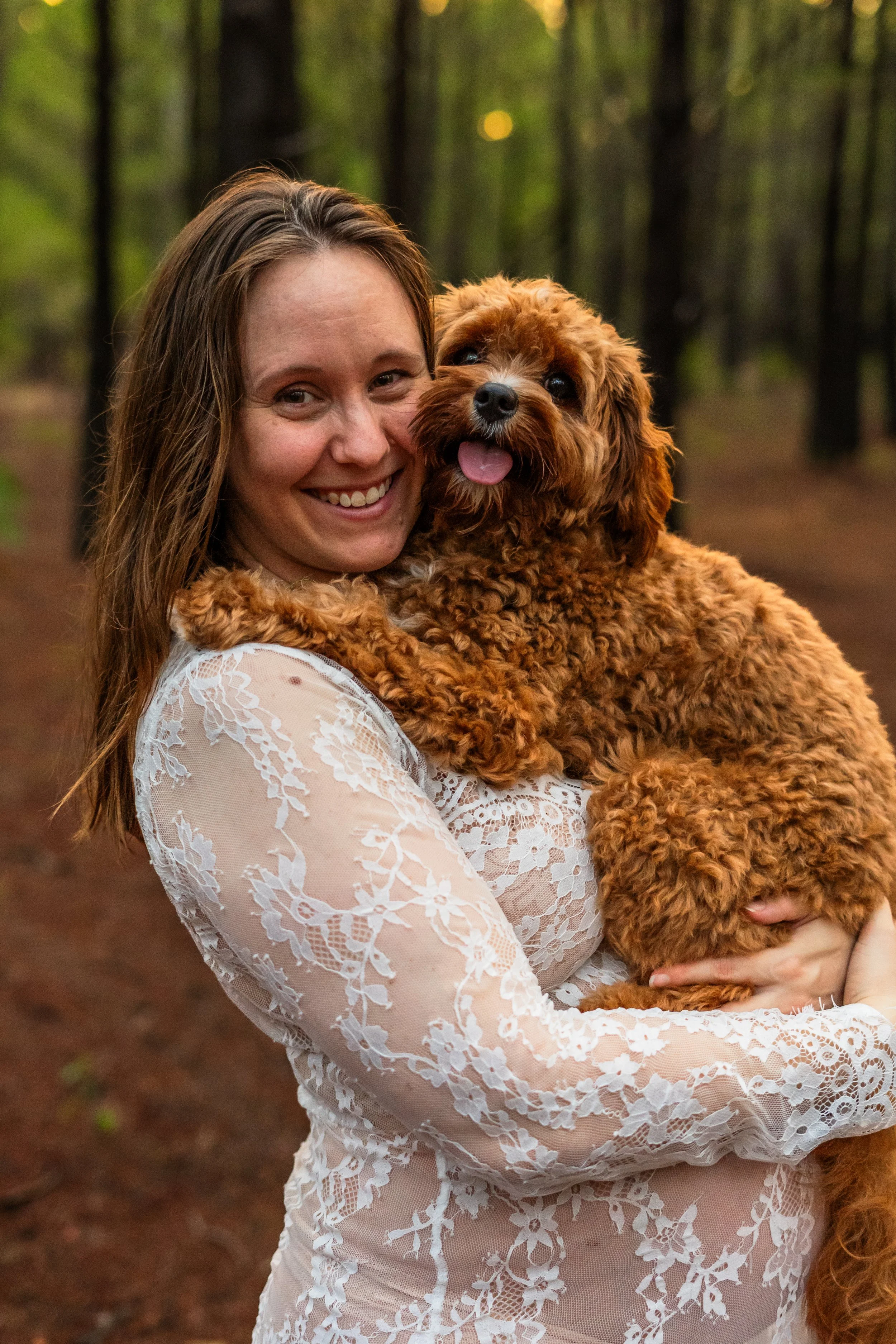 A woman holding a fluffy brown dog with a curly coat in a forested area during daylight.