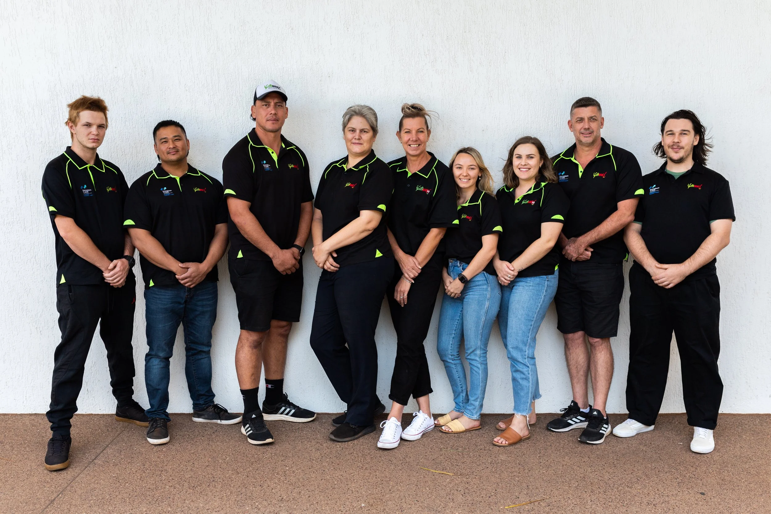 Group of nine people standing in front of a white wall, wearing black shirts with green accents, posing for a team photo.