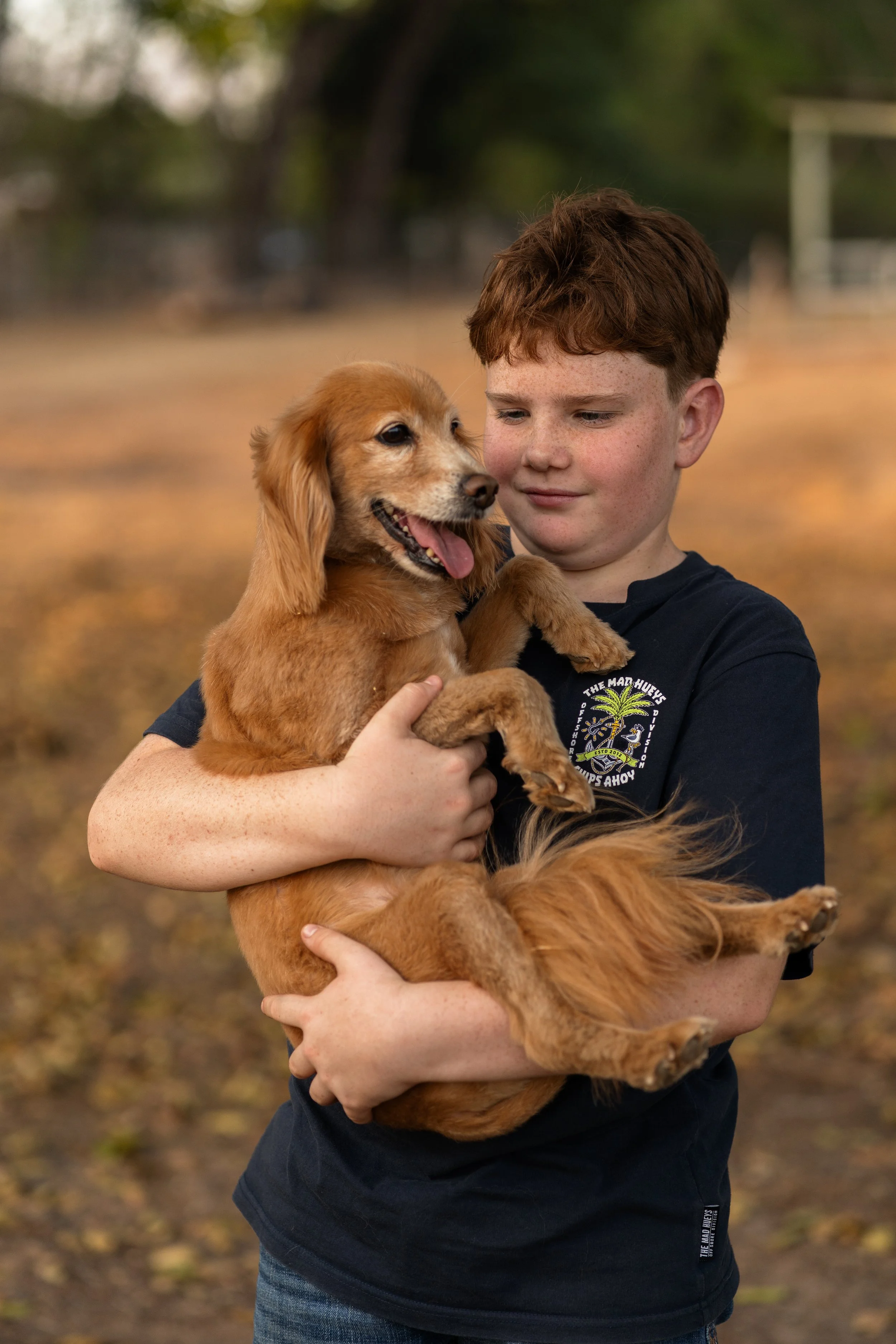 A young boy with red hair holding a golden retriever puppy outdoors. The background is blurred with trees and fallen leaves.