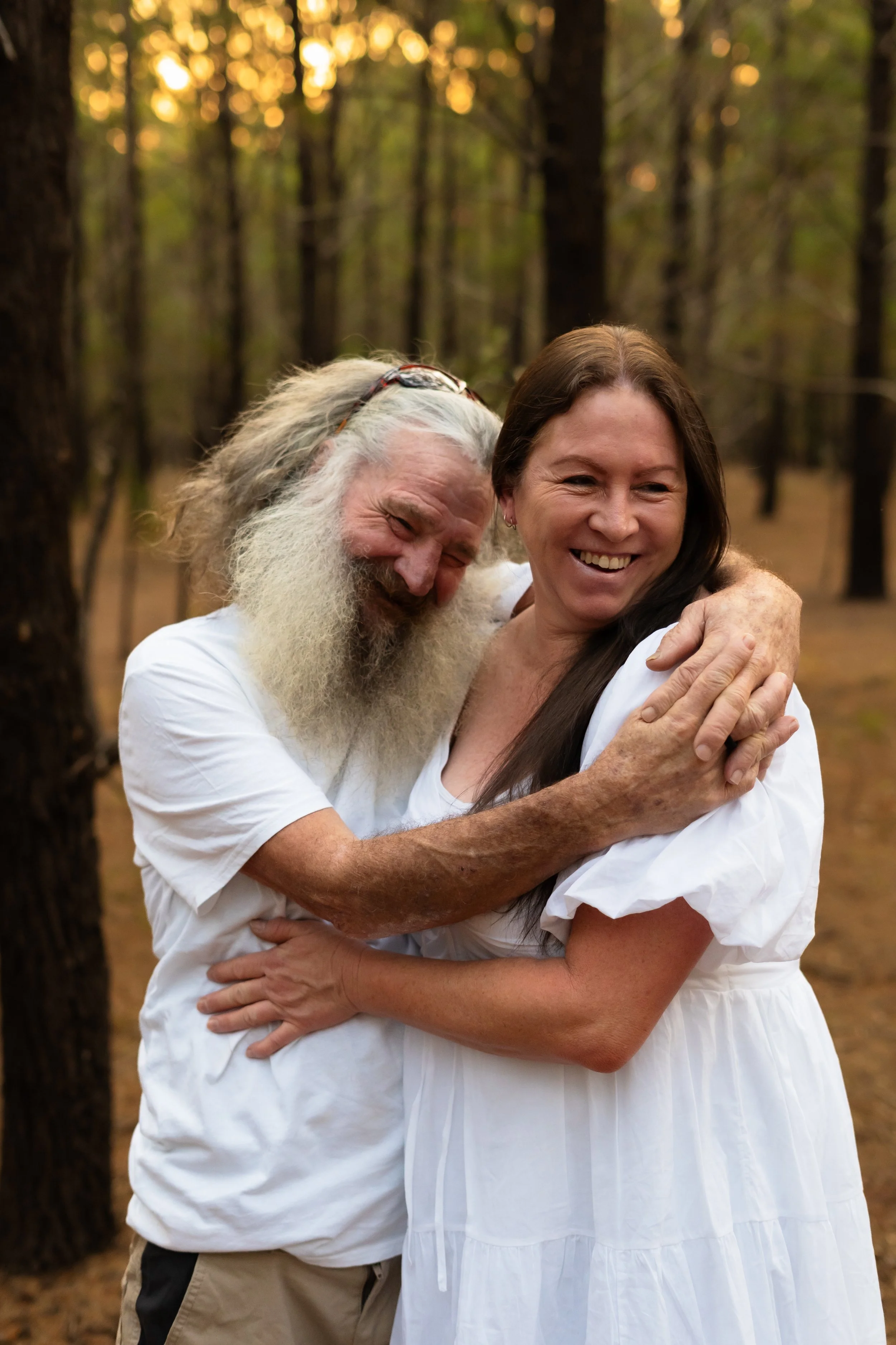 An elderly man with long gray hair and a beard hugging a smiling woman with long brown hair in a white dress, standing in a wooded area during sunset.