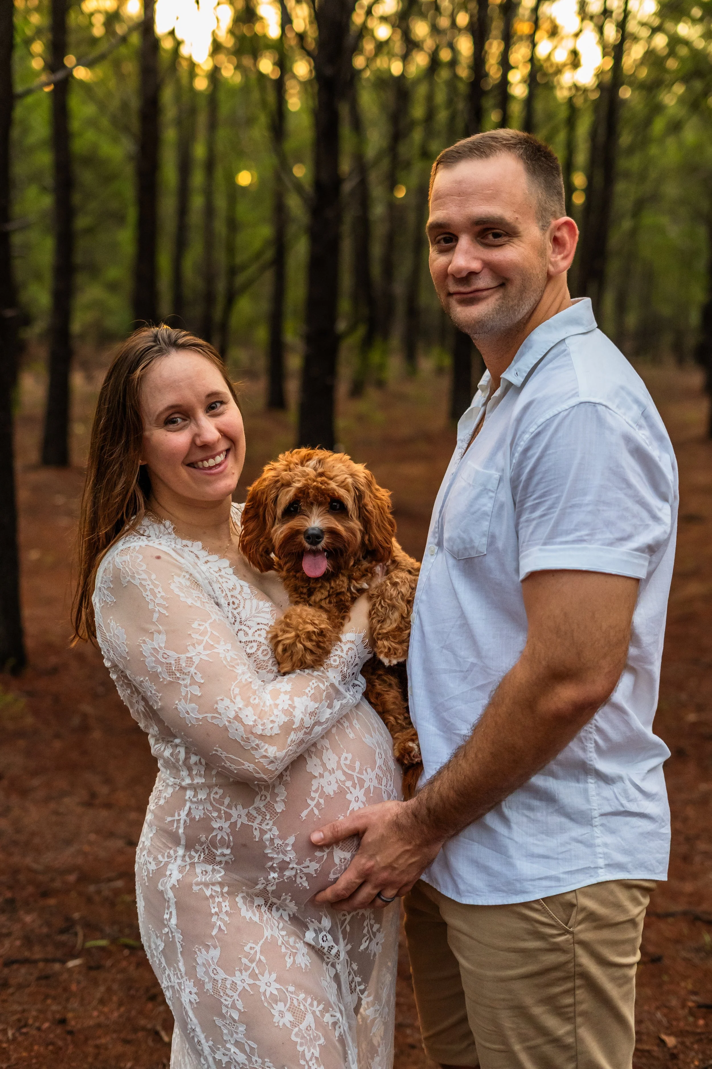 A pregnant woman in a white lace dress holding a brown puppy, standing beside a man in a light blue shirt in a wooded area during sunset.