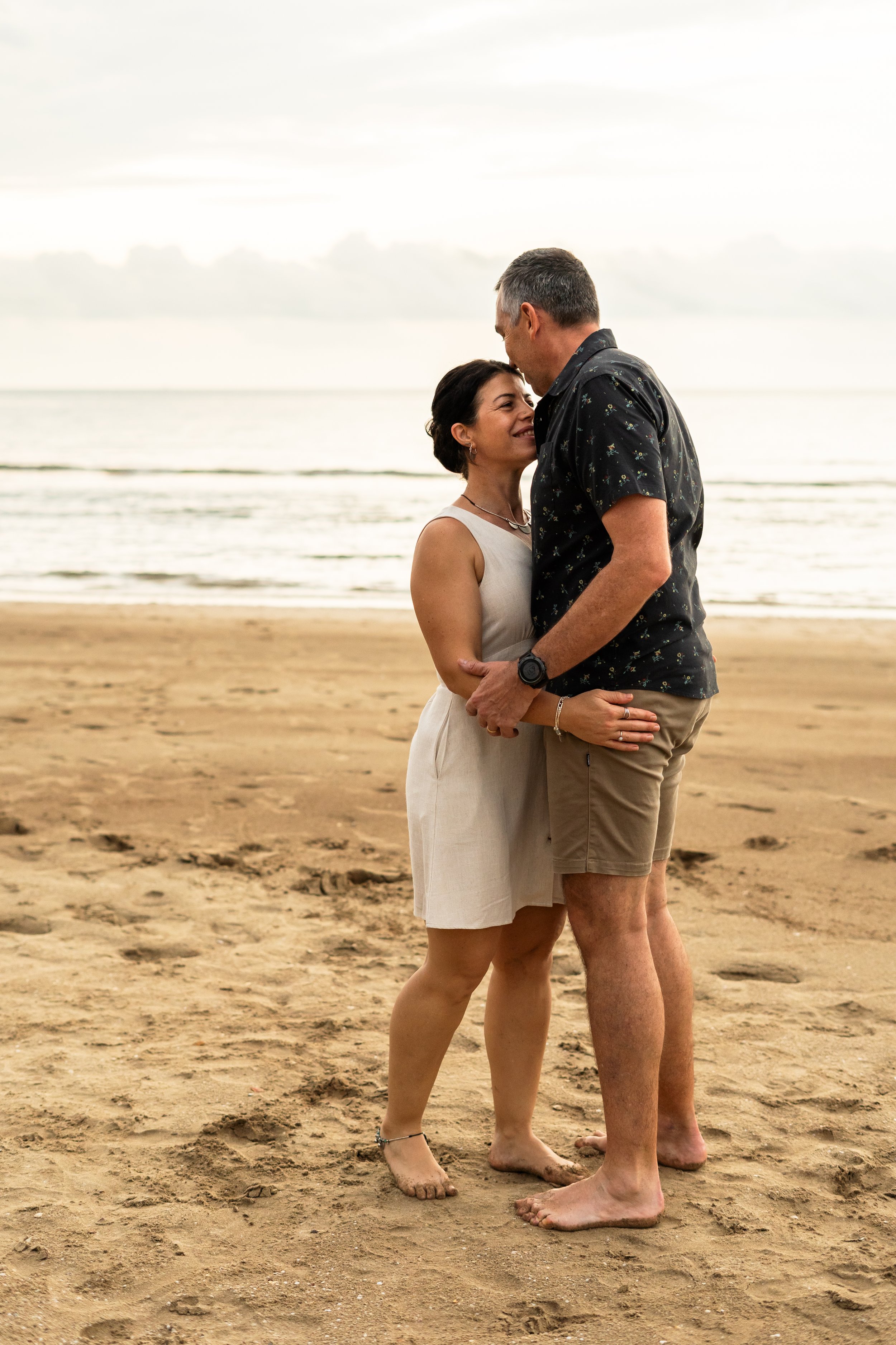 A couple standing close on a sandy beach during sunset, gazing at each other with the ocean and cloudy sky in the background.