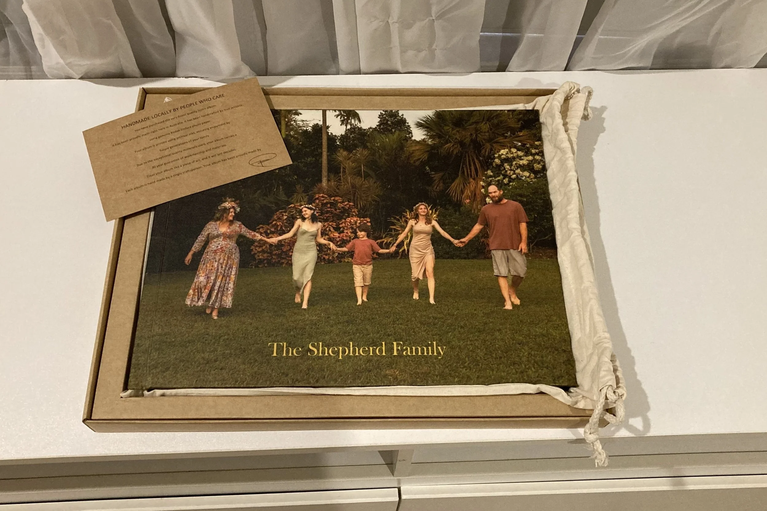 A customized photo album titled 'The Shepherd Family' with a photo of five people holding hands and walking on grass, surrounded by trees. The album is inside a beige cardboard box with a cloth lining. A small brown card with text is placed on top of